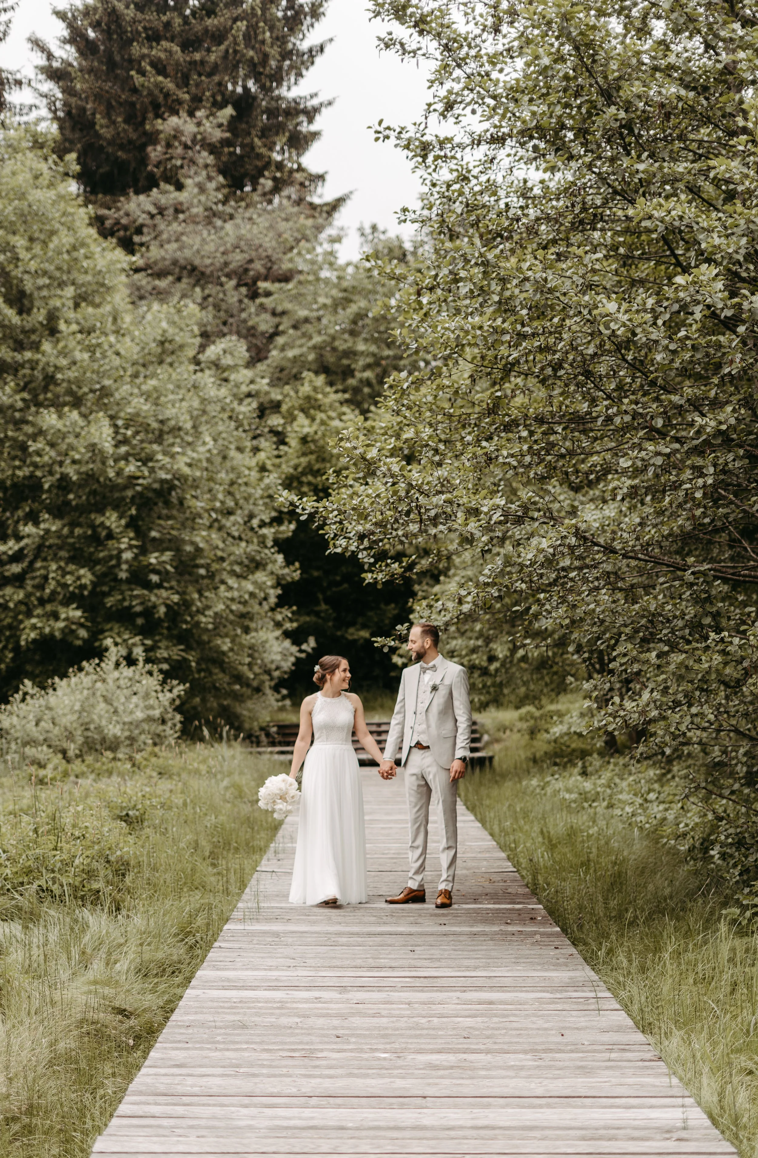 Ein frisch vermähltes Paar hält Händchen auf einem Holzsteg in einer grünen, bewaldeten Umgebung.
