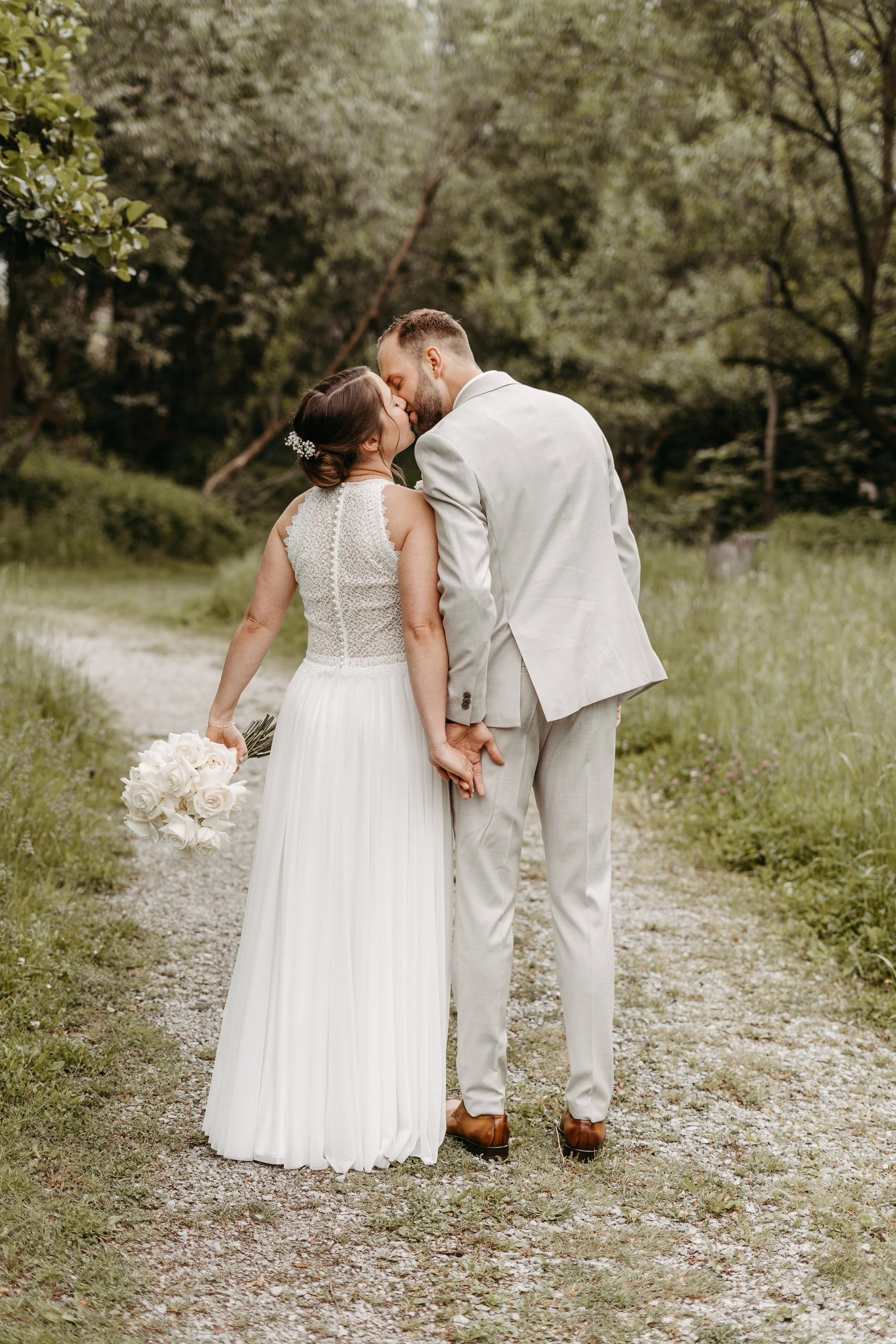 Ein Brautpaar in Hochzeitskleidung küsst sich auf einem Waldweg in der Natur, die Braut hält einen weißen Blumenstrauß in der Hand.