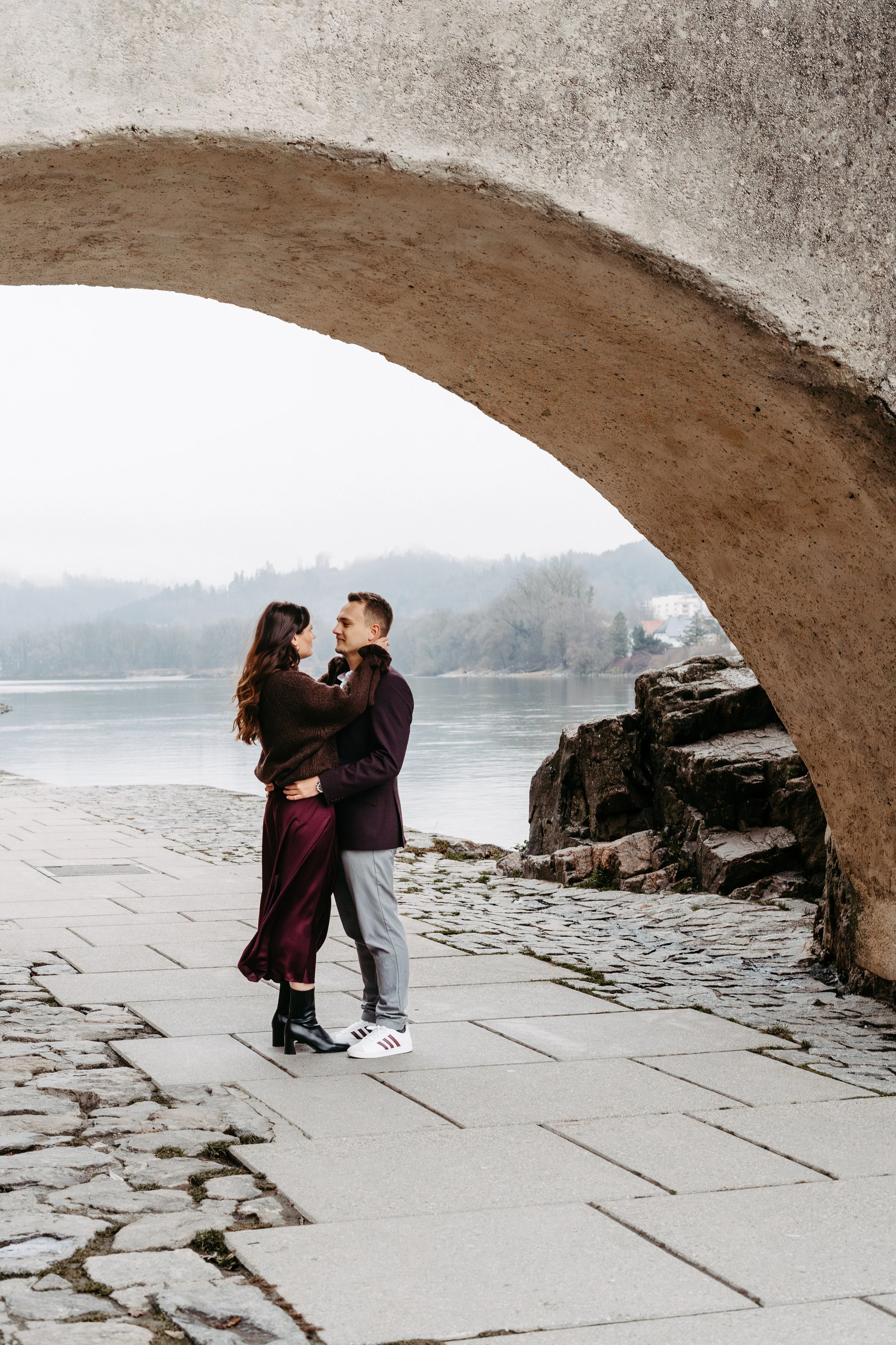 Ein Paar steht sich unter einer Brücke am Flussufer, umarmt sich liebevoll, im Hintergrund sind Bäume und ein nebliger Himmel zu sehen.
