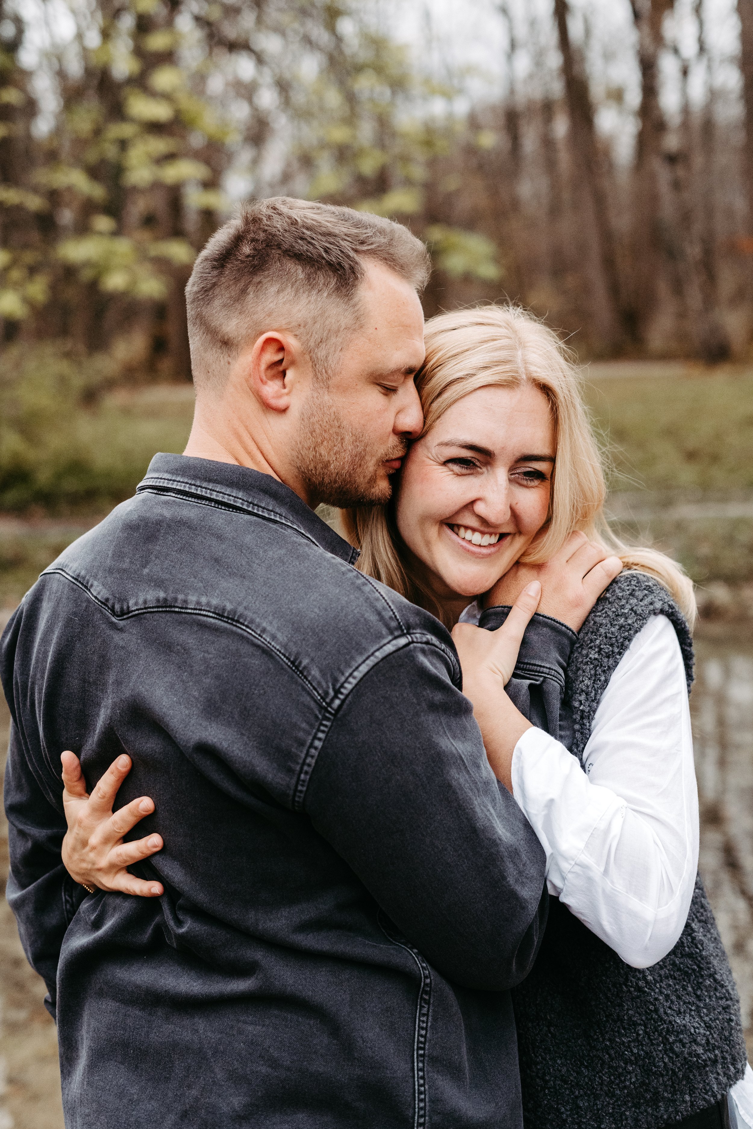 Ein Paar umarmt sich liebevoll in der Natur an einem bewölkten Tag, im Hintergrund Bäume und Wasser.