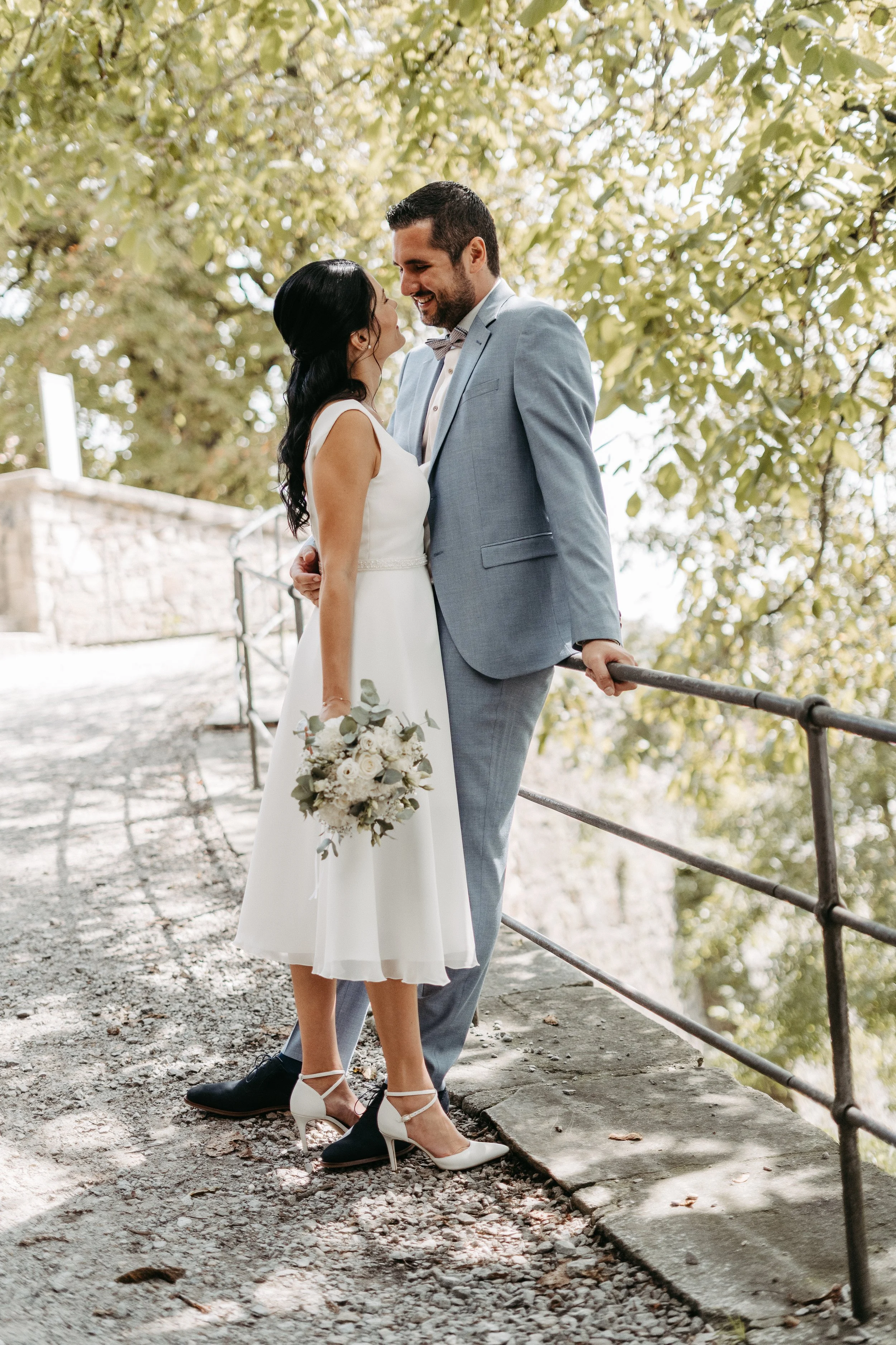 Ein Brautpaar steht sich auf einer Brücke im Freien gegenüber, umgeben von grünen Bäumen, sie lächeln sich an und halten sich fest.