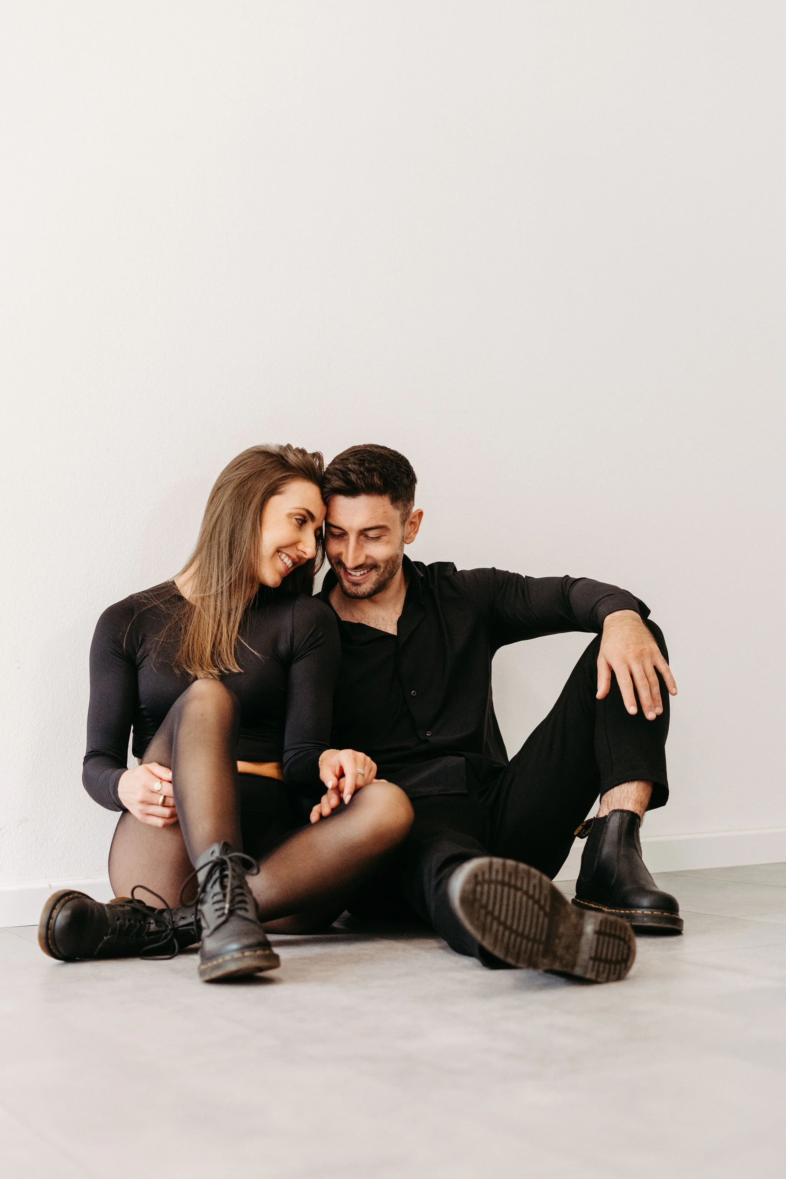 Ein lachendes Paar sitzt auf dem Boden vor einer weißen Wand, Frau trägt schwarze Kleidung, Mann trägt schwarze Kleidung und Stiefel.