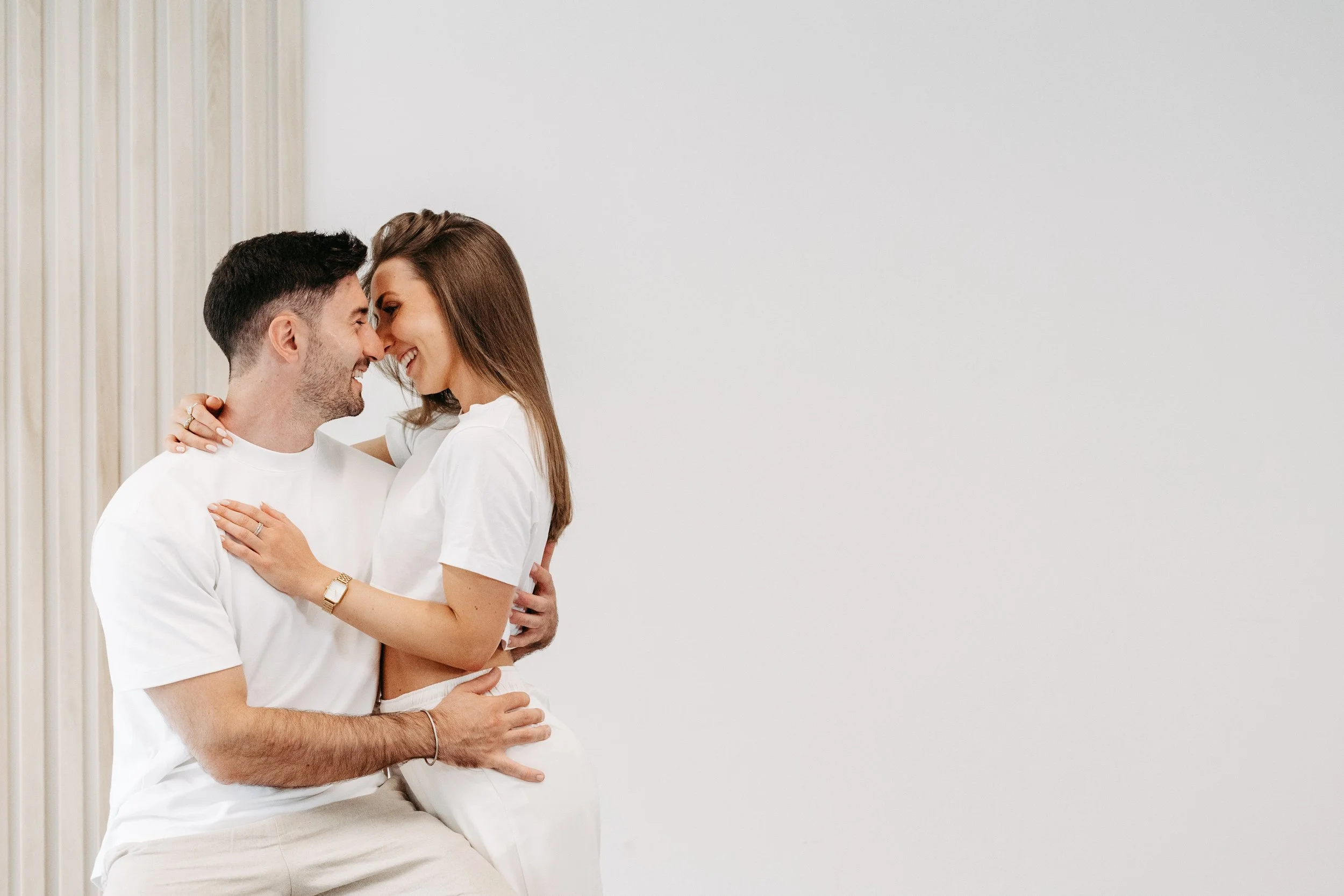 Ein glückliches Paar, das sich liebevoll umarmt und lacht, in einem minimalistischen Raum mit weißen Wänden und Vorhängen.