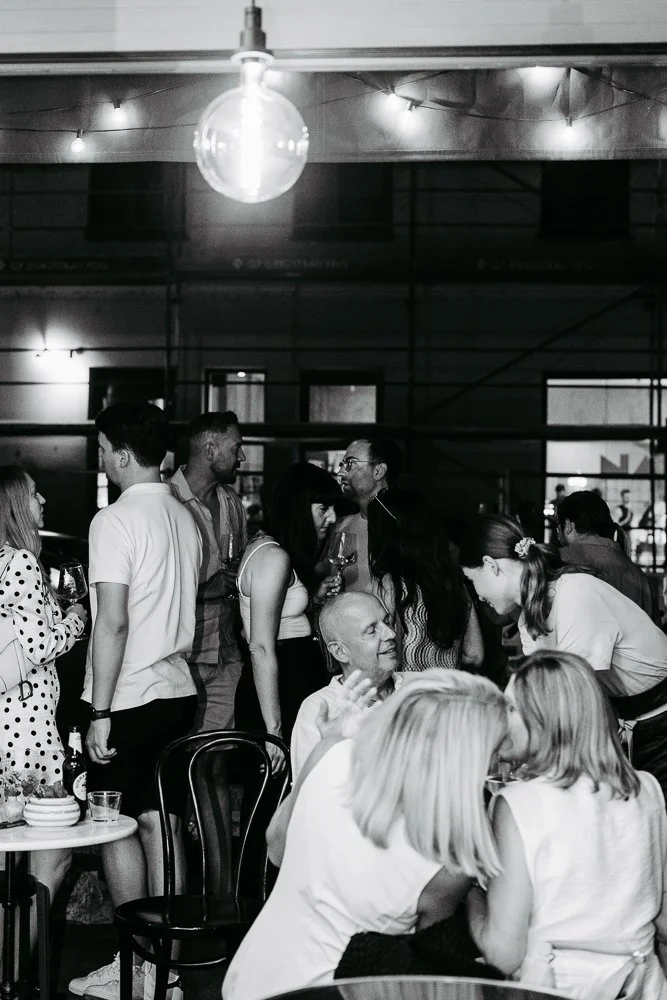 Mehrere Menschen beim Zusammenkommen in einem Raum, einige sitzen an Tischen, andere stehen und sprechen, mit Gläsern in der Hand.