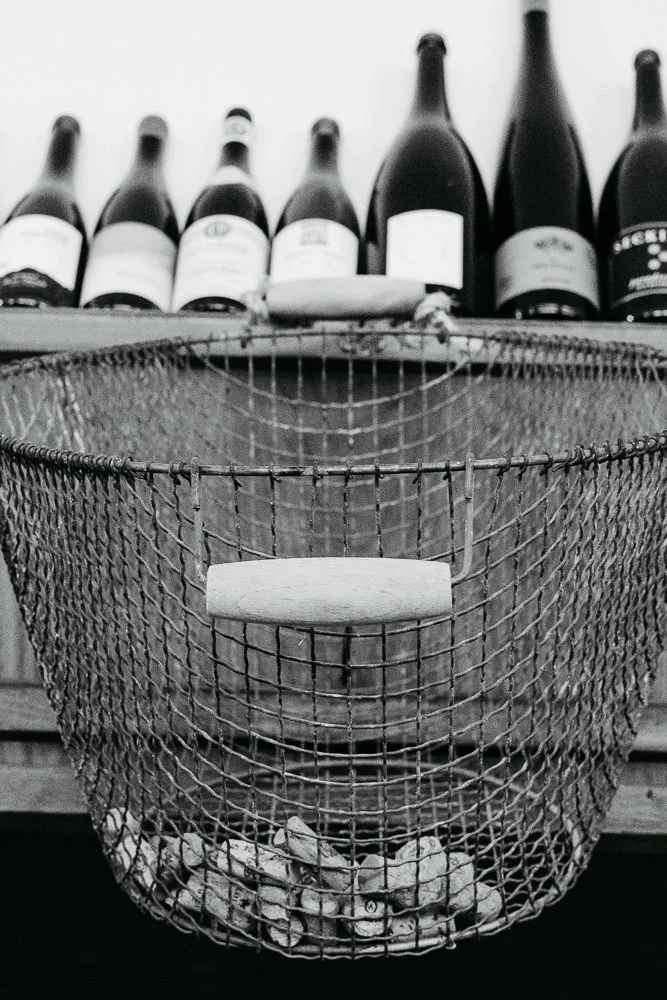 Ein leeres Metalldrahtkorb mit Holzgriff, darunter Steine, im Hintergrund Weinflaschen auf einer oberen Ablage