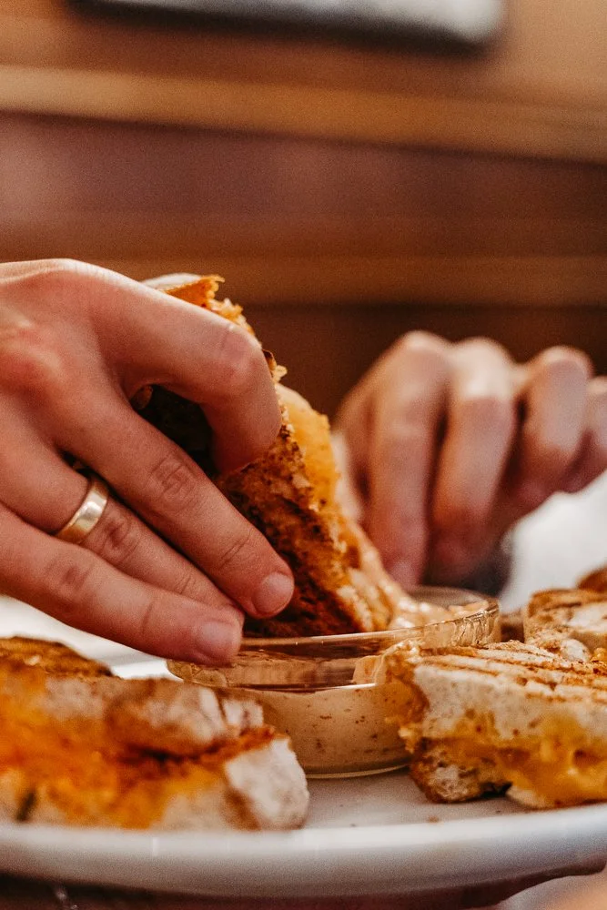 Person, die ein Sandwich mit Käse isst, mit einem Teller voller Sandwiches im Vordergrund.
