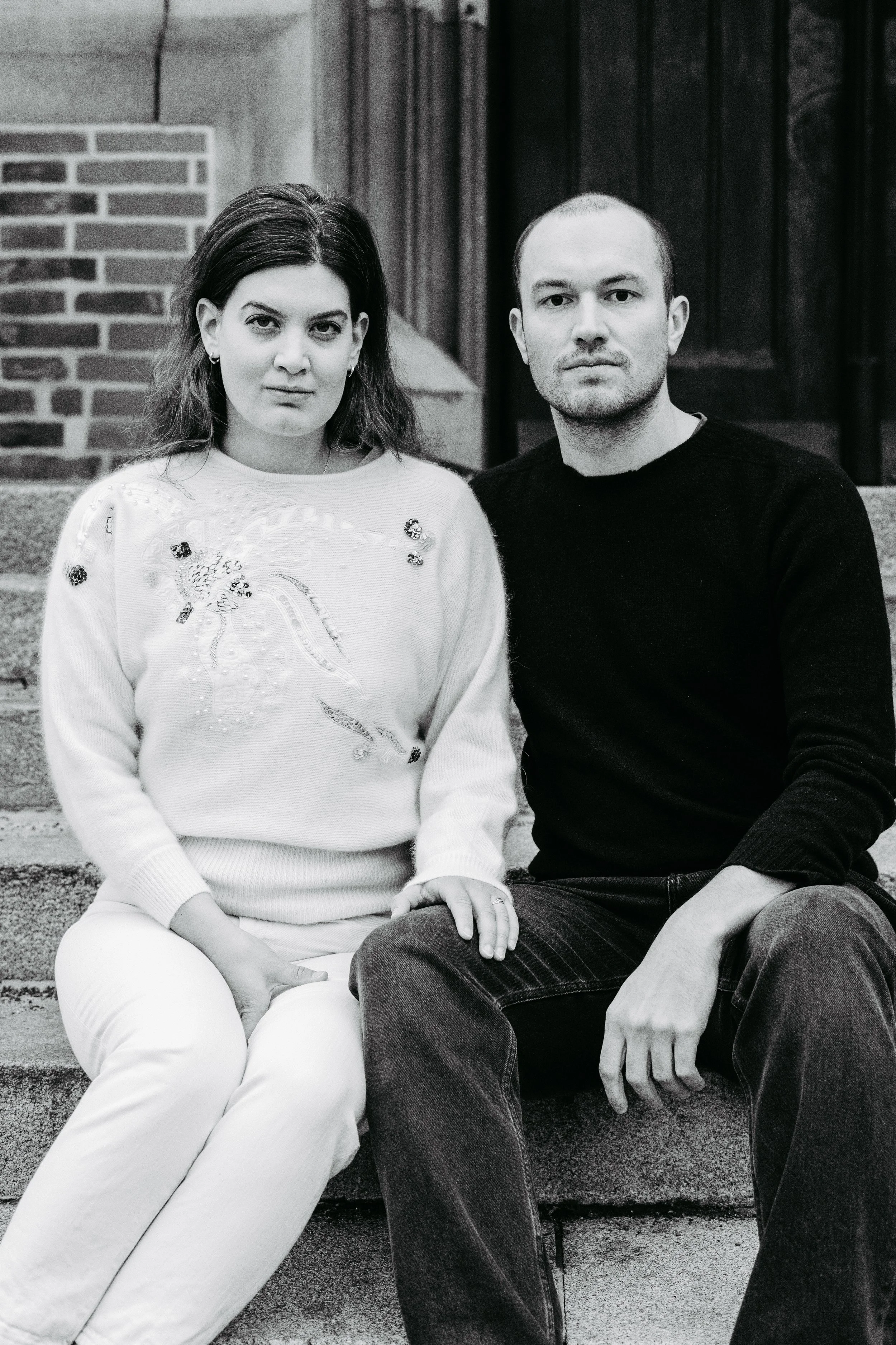 Ein Mann und eine Frau sitzen nebeneinander auf Steinstufen vor einem Gebäude, Schwarz-Weiß Foto.