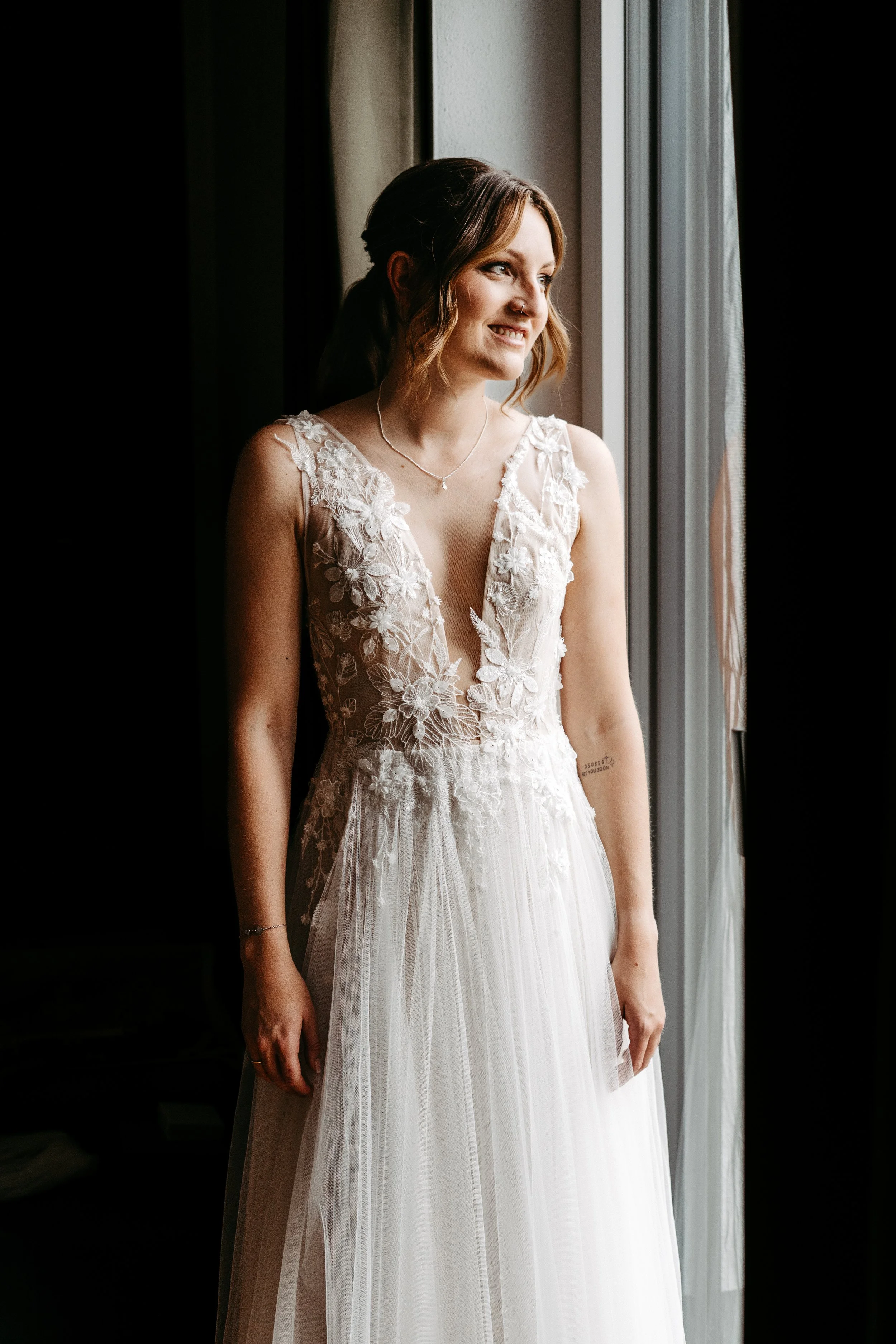 Braut in Hochzeitskleid steht am Fenster und lächelt.