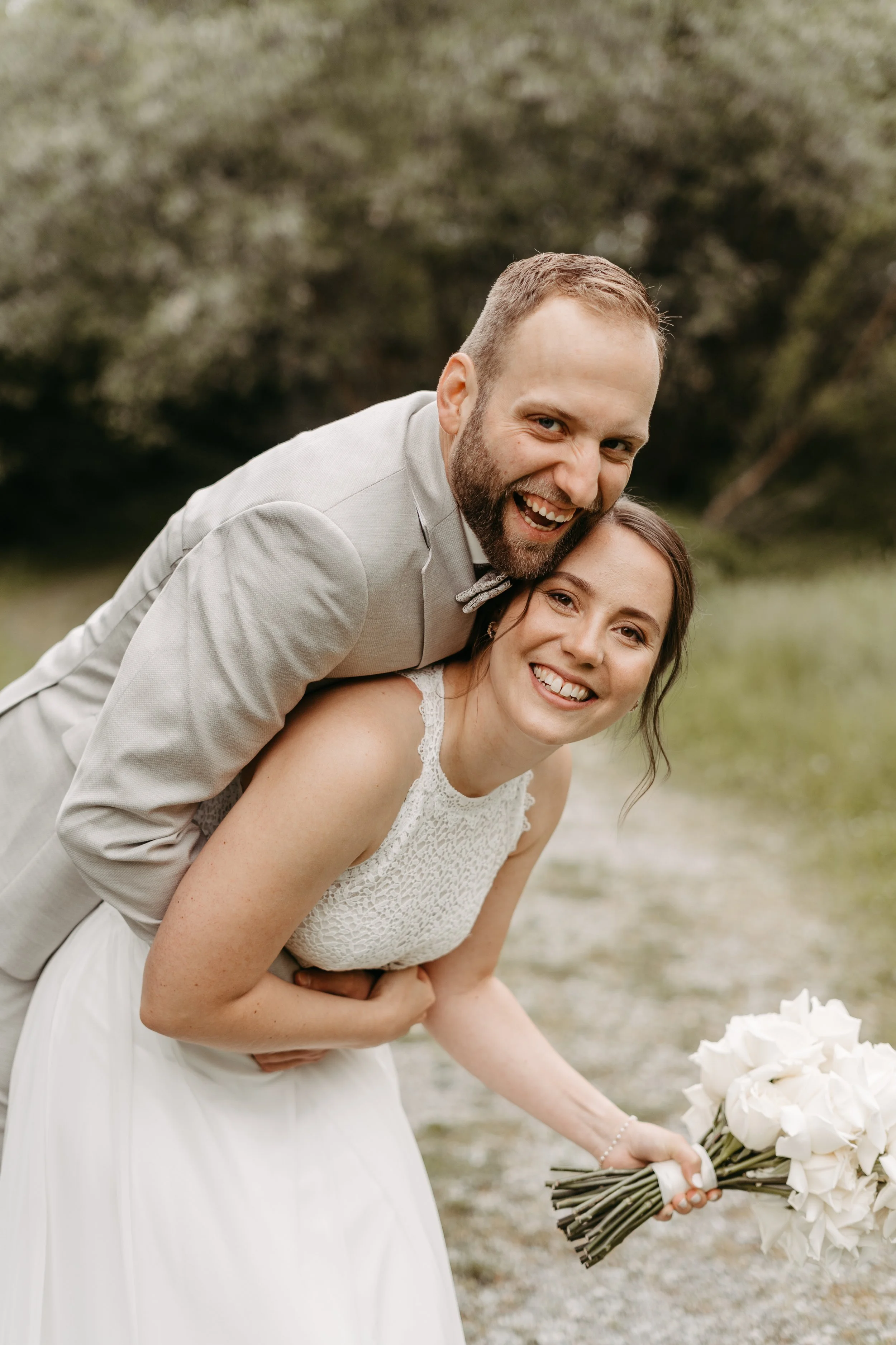 Ein lachendes Brautpaar bei einer Hochzeit im Freien, der Bräutigam trägt einen hellgrauen Anzug und die Braut hält einen weißen Blumenstrauß.