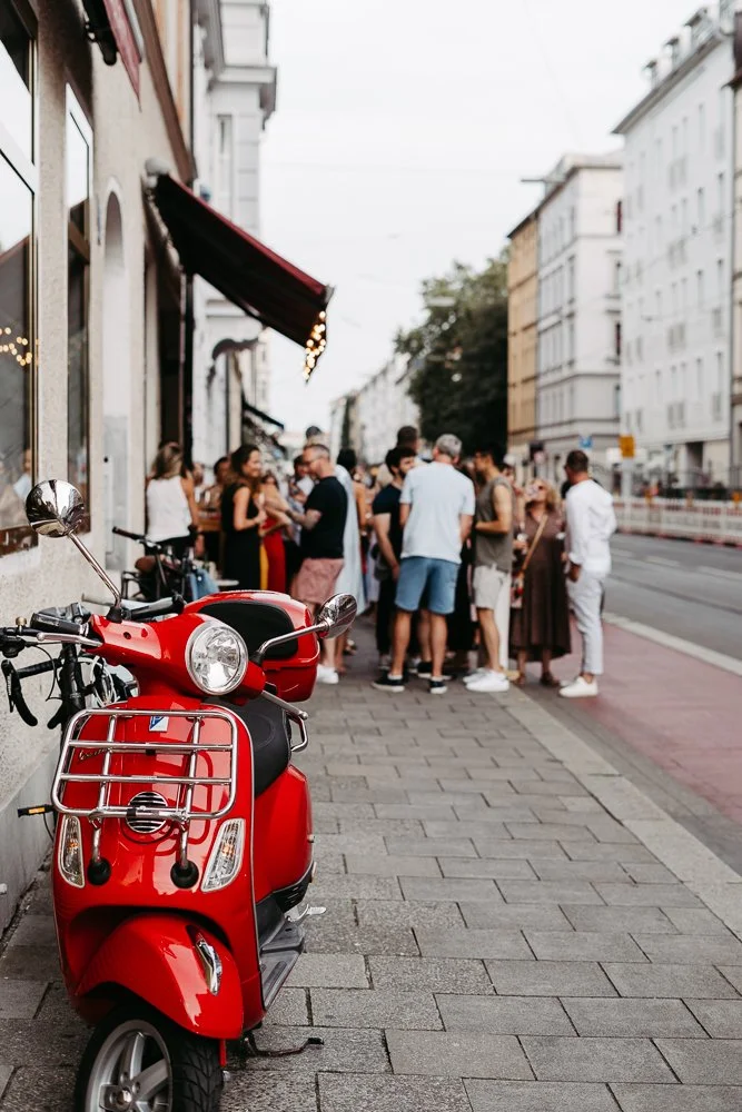 Eine rote Vespa steht an der Straßenseite, während eine Gruppe von Menschen draußen vor einem Geschäft auf dem Bürgersteig versammelt ist.