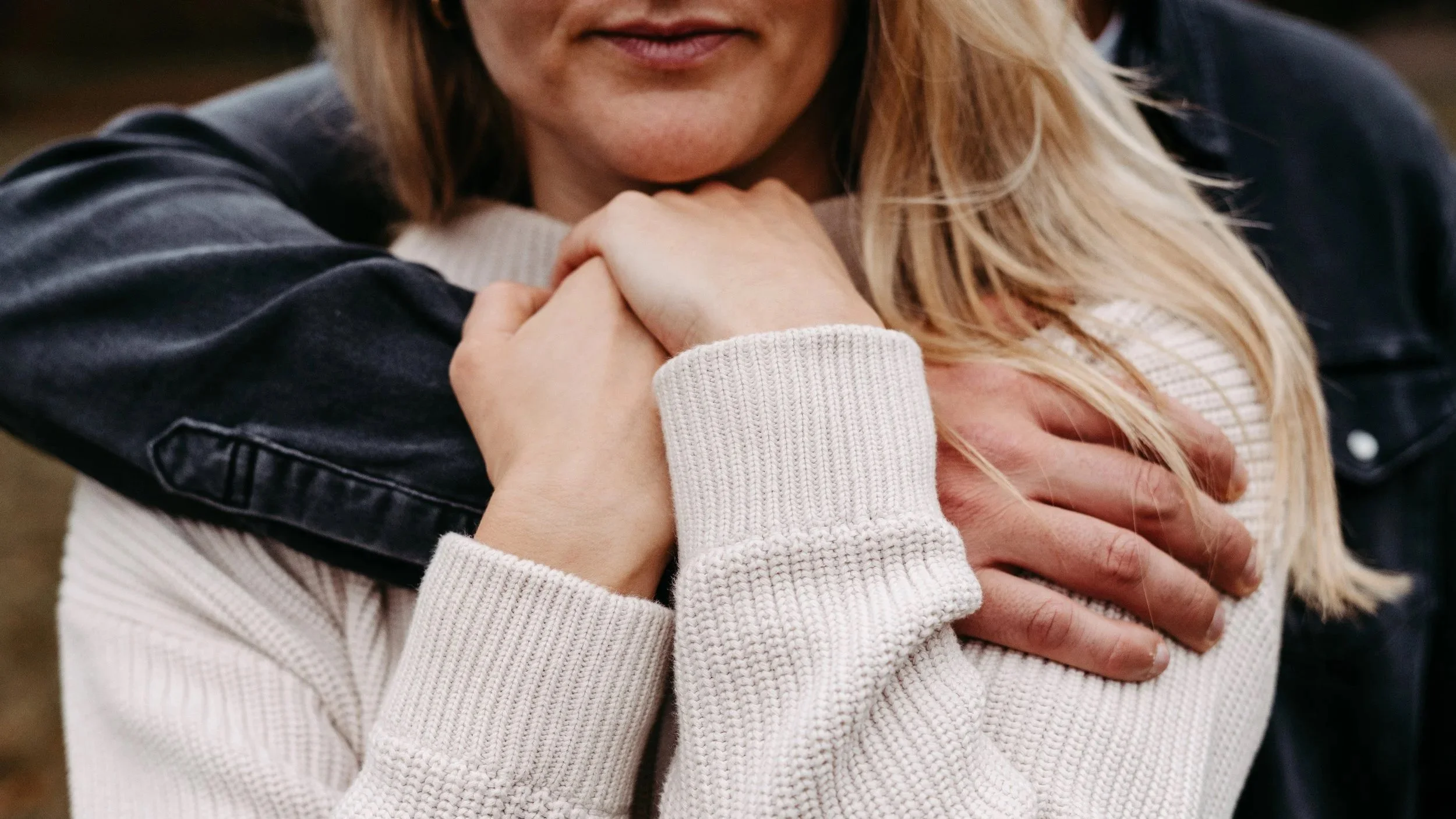 Zwei Menschen umarmen sich, eine Person trägt ein weißes Strickshirt und eine schwarze Jacke, die andere hat eine dunkle Jacke.