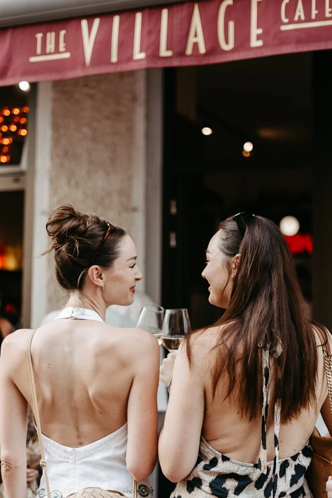 Zwei Frauen trinken Wein und sprechen in einem Restaurant oder einer Bar, im Hintergrund ein Schild mit der Aufschrift 'The Village'.