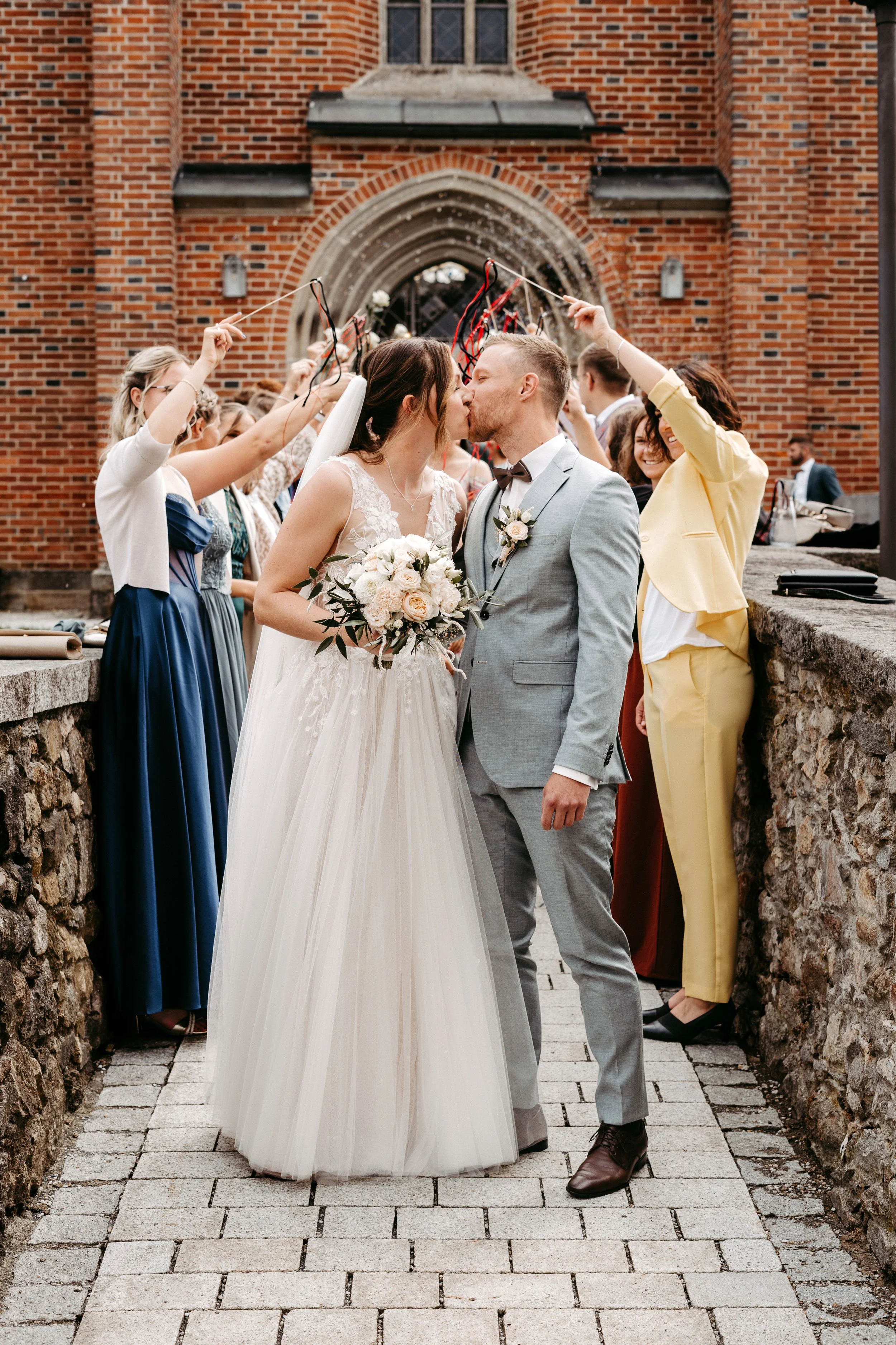 Ein brautpaar küsst sich vor einer Gruppe von Freunden vor einer roten Backstein-Kirche. Die Freunde halten Luftballons hoch, um das Brautpaar zu feiern.
