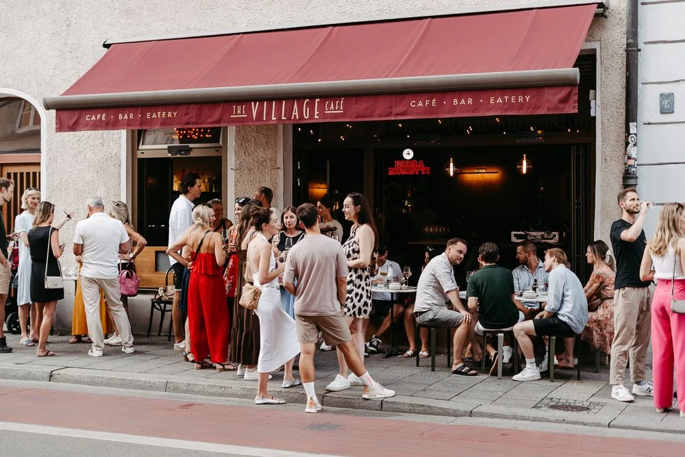 Menschen vor einem Café mit rotem Sonnenschutz, einige sitzen an Tischen, andere stehen in der Warteschlange, bei Nacht.