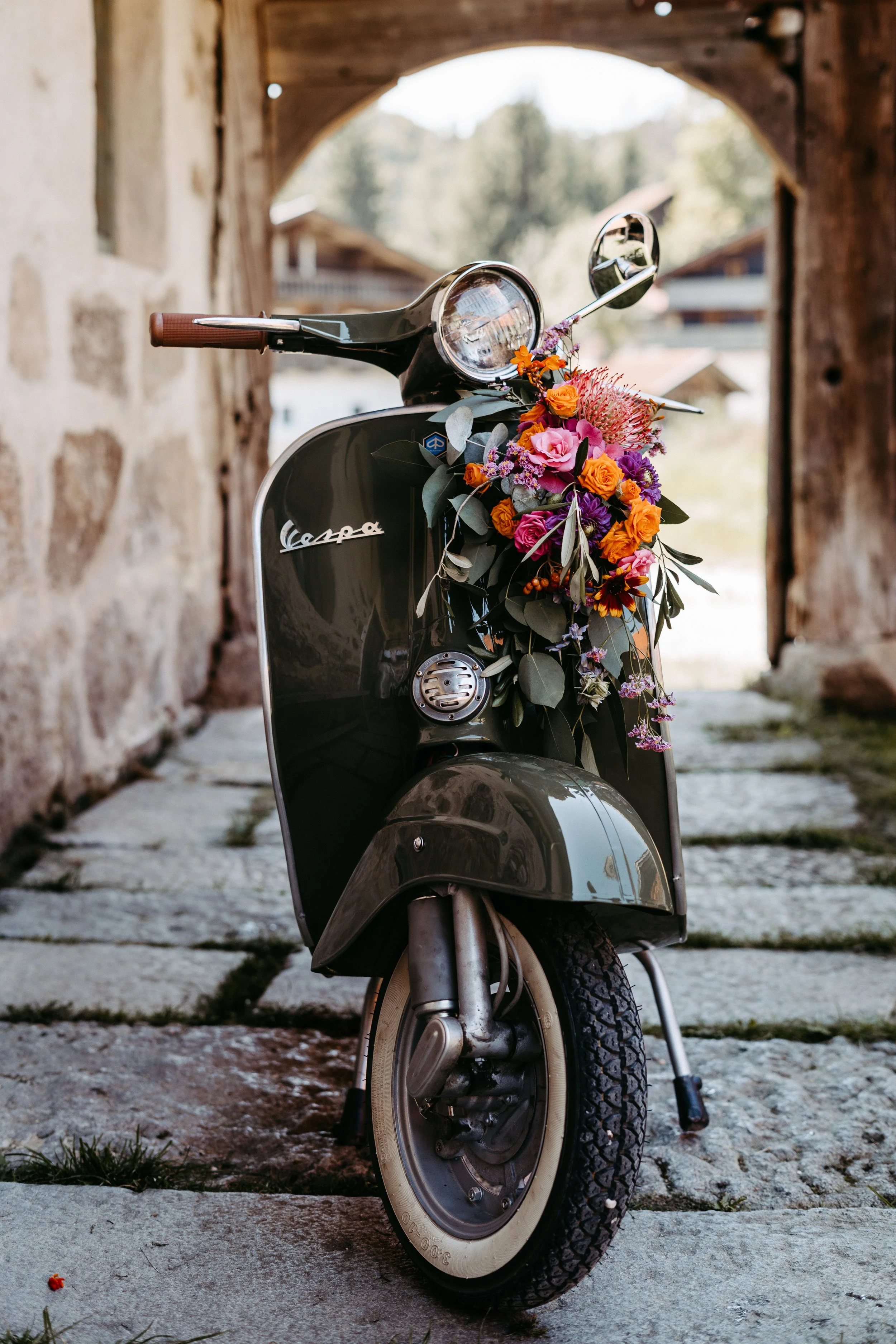 Ein schwarzer Vespa-Roller mit einem Blumenstrauß an der Vorderseite, steht vor einer Steinmauer und einem Holztor, im Hintergrund Bäume und ein Haus.