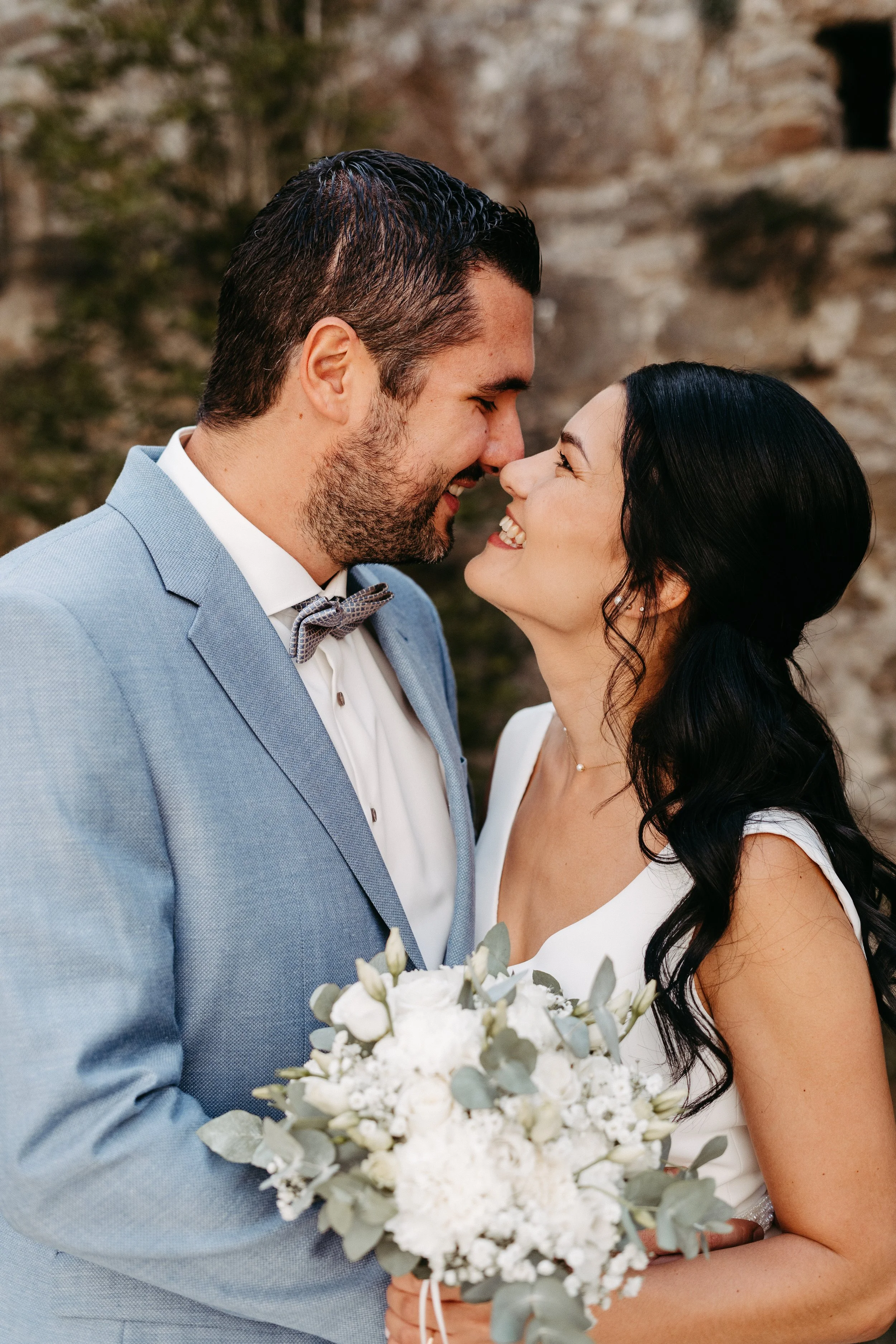 Ein glückliches Paar bei einer Hochzeit, das sich liebevoll gegenübersitzt und in die Augen schaut, mit einem Brautstrauß in der Hand, vor einer Naturkulisse im Freien.