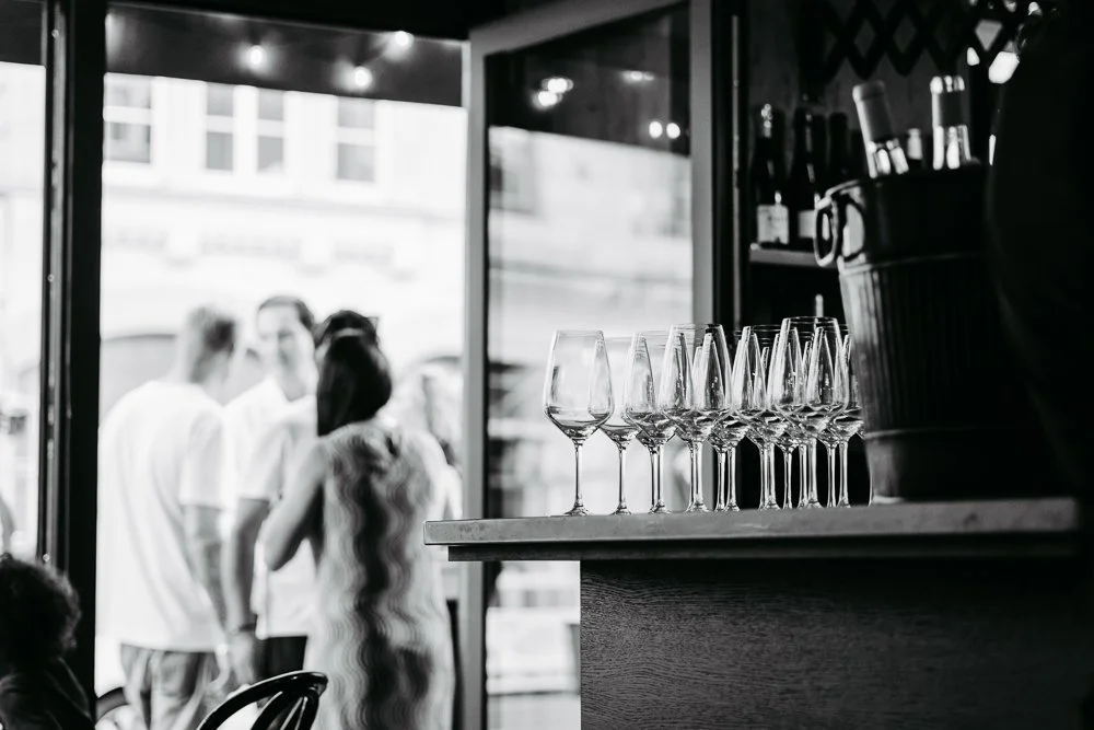 Leere Weingläser auf einem Tisch in einem Café, im Hintergrund Menschen beim Gespräch, Blick nach draußen durch große Fenster