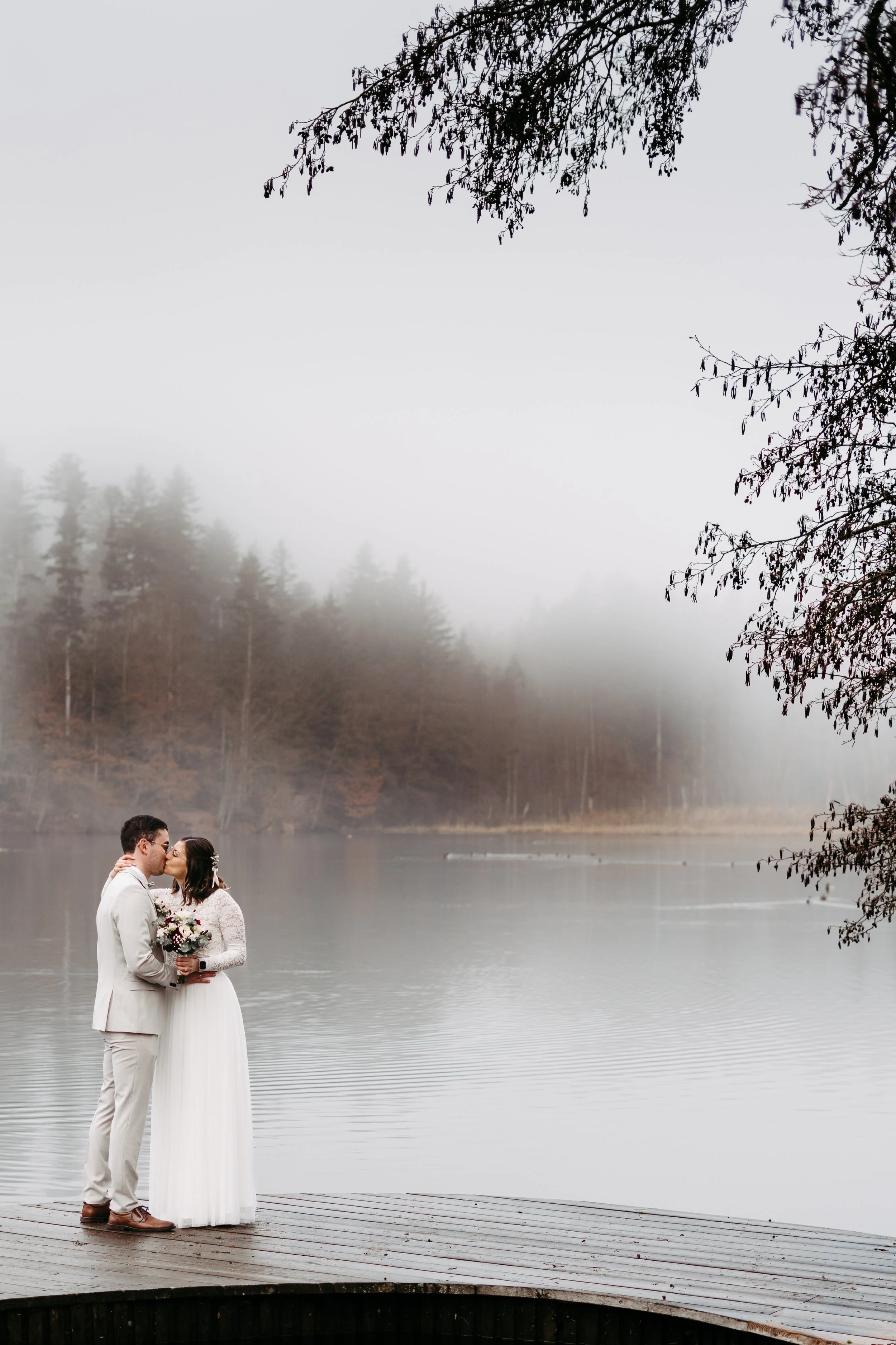 Ein Hochzeitsfoto von einem Paar, das sich an einem nebligen See küsst, mit Bäumen im Hintergrund und zweigen im Vordergrund.