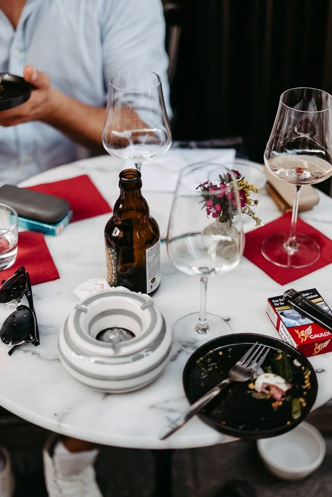 Leere Wein- und Biergläser auf einem weißen Marmortisch, zerdrücktes Teller mit Essensresten, Blume in Vase, offene Zigarettenschachtel, Handy, Sonnenbrille, und leere Flasche.