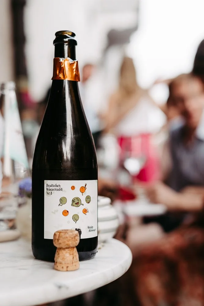 Eine Flasche mit einem Etikett und ein Korken auf einem Tisch, im Hintergrund Menschen, die zusammen feiern oder essen.