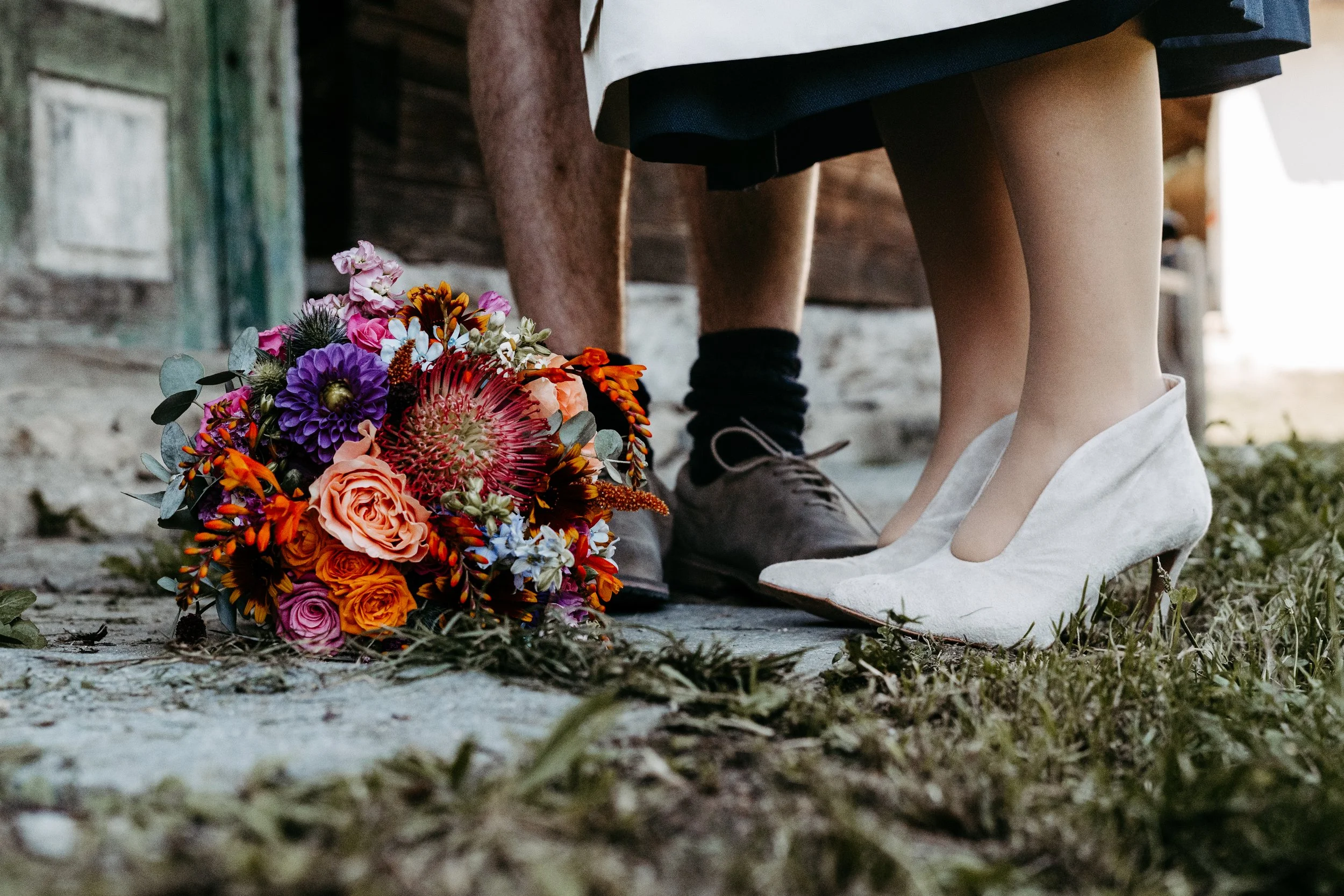 Ein Brautpaar steht mit den Füßen auf einem Kopfsteinpflaster, das Brautpaar trägt weiße High Heels, und zwischen ihren Füßen liegt ein bunter Blumenstrauß auf dem Boden.