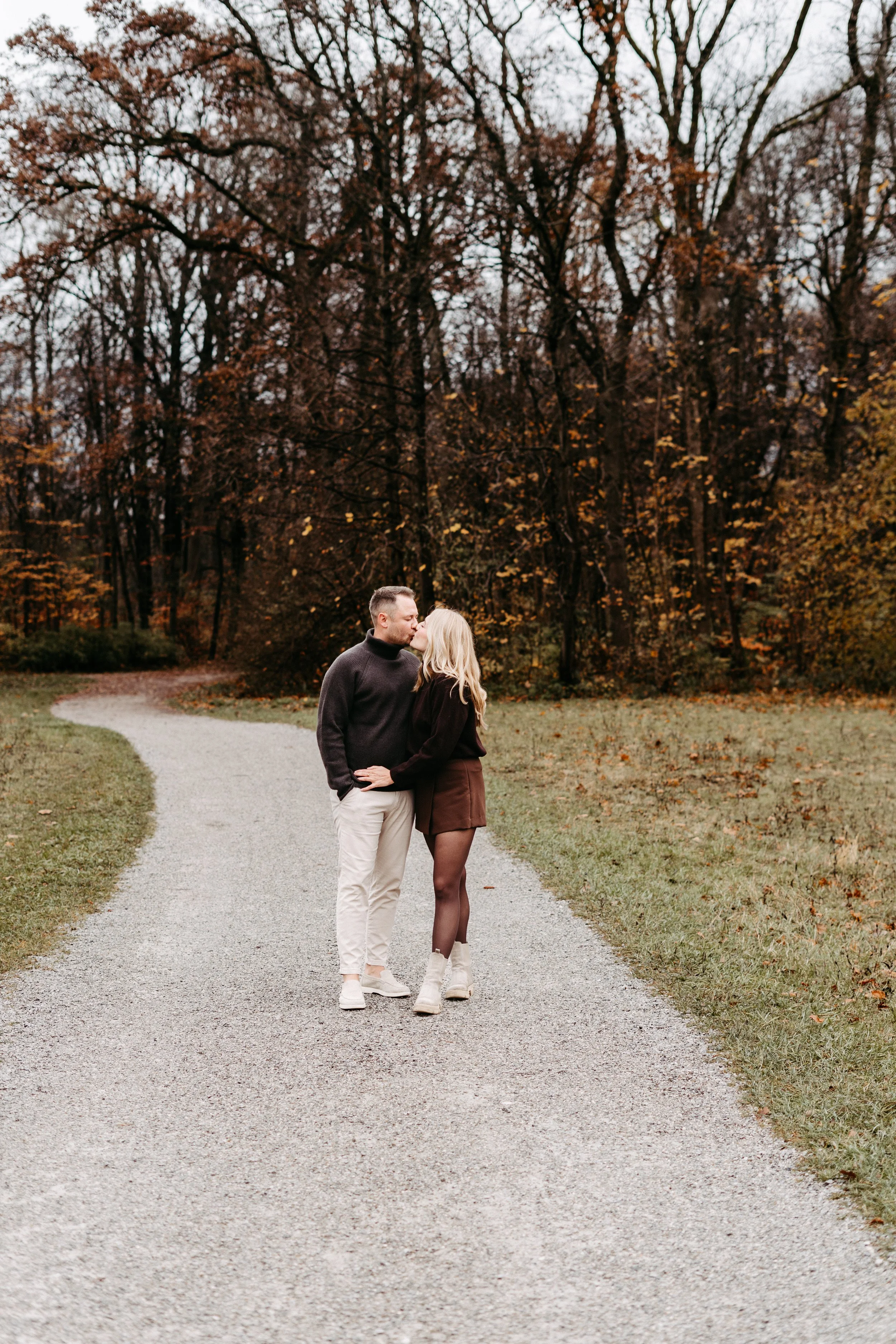 Ein Paar küsst sich auf einem Weg durch einen Herbstwald, Bäume mit braunen Blättern im Hintergrund.