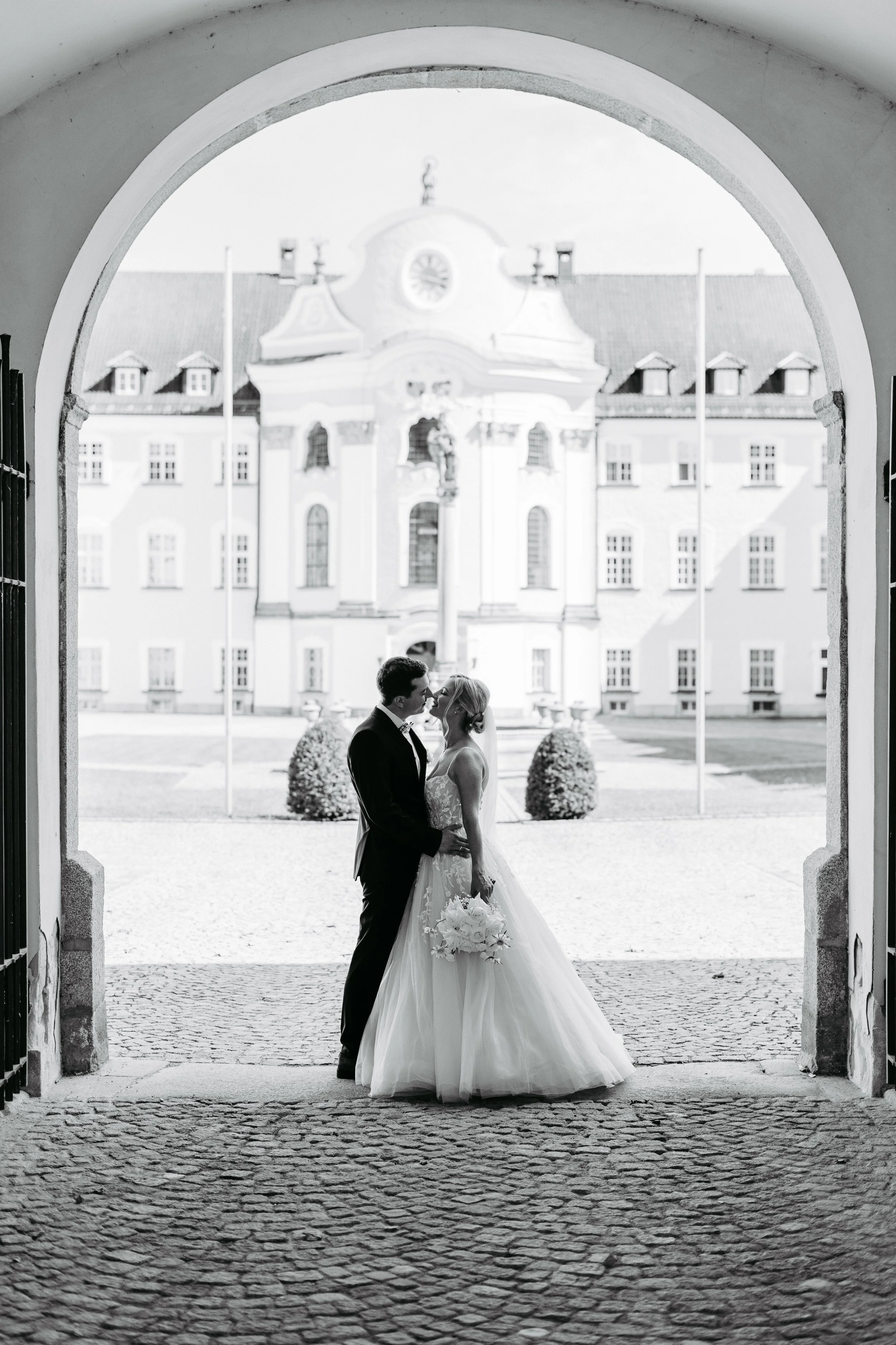 Ein Brautpaar steht sich bei ihrer Hochzeit unter einem Torbogen gegenüber, mit einem historischen Gebäude im Hintergrund.