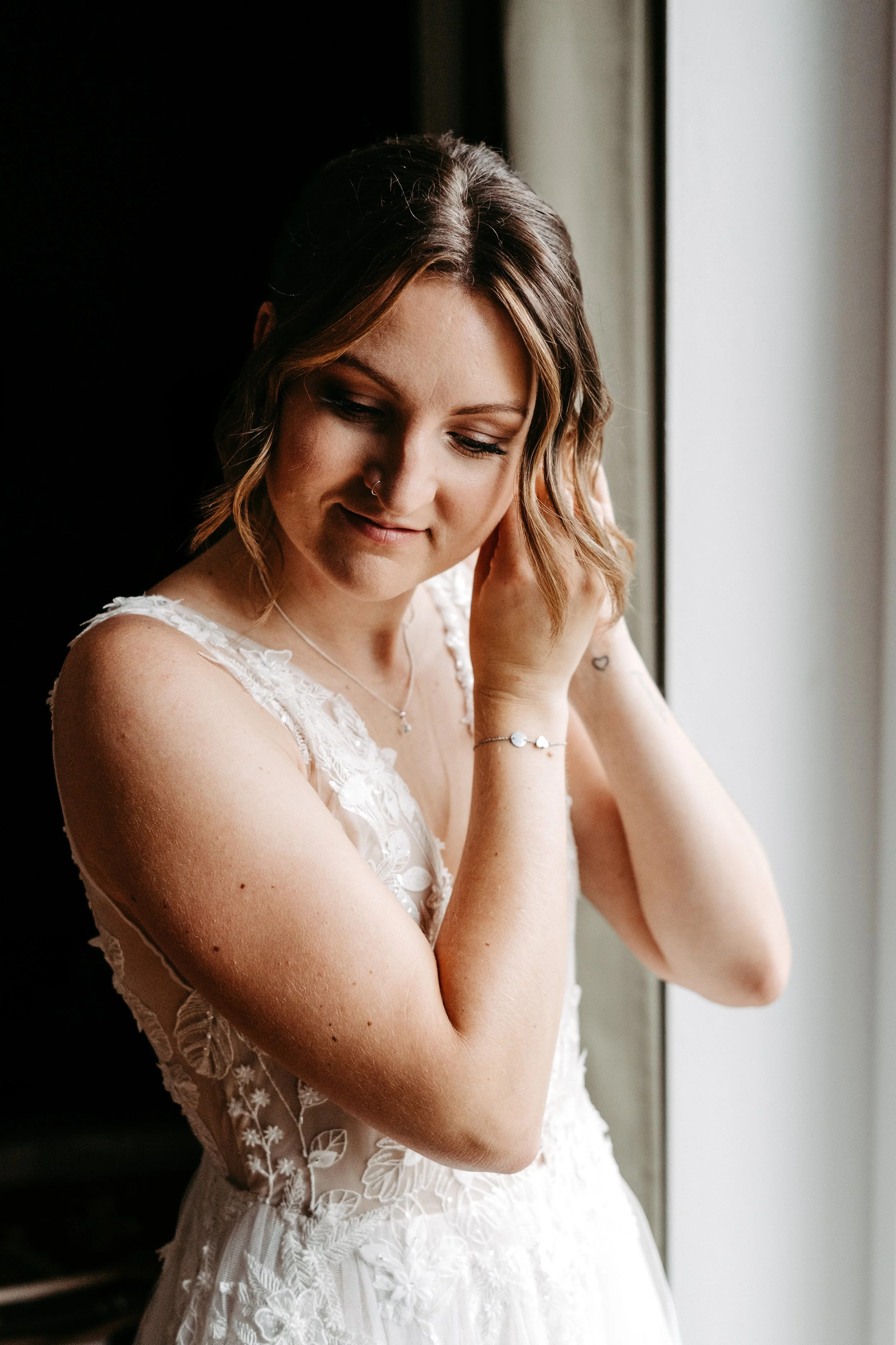 Eine junge Frau in einem weißen, eleganten Hochzeitskleid steht neben einem Fenster, berührt ihr Ohr und lächelt leicht.