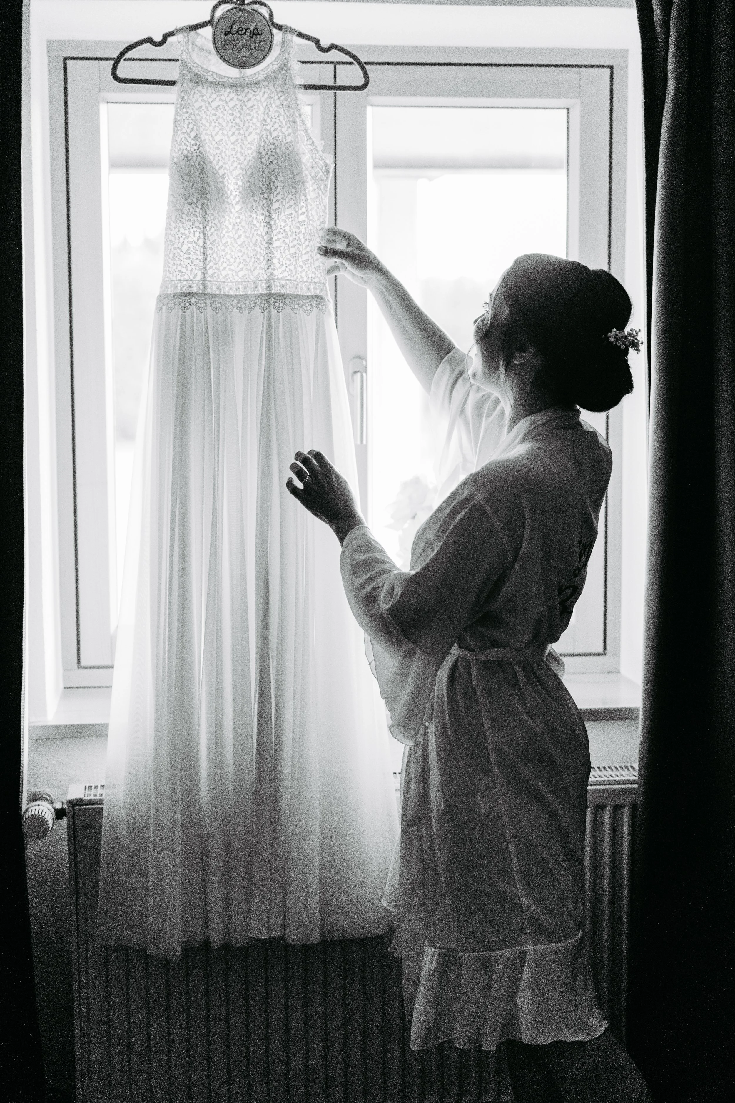 Eine Frau betrachtet ein Hochzeitskleid, das an einem Kleiderbügel vor einem Fenster hängt.