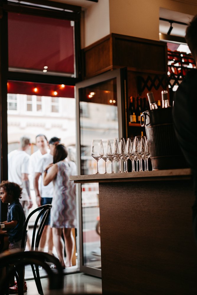 Innenaufnahme eines Cafés oder Restaurants mit leer gestellten Gläsern auf der Theke und einigen Personen im Hintergrund, die sich vor einem geöffneten Fenster oder Tür unterhalten.