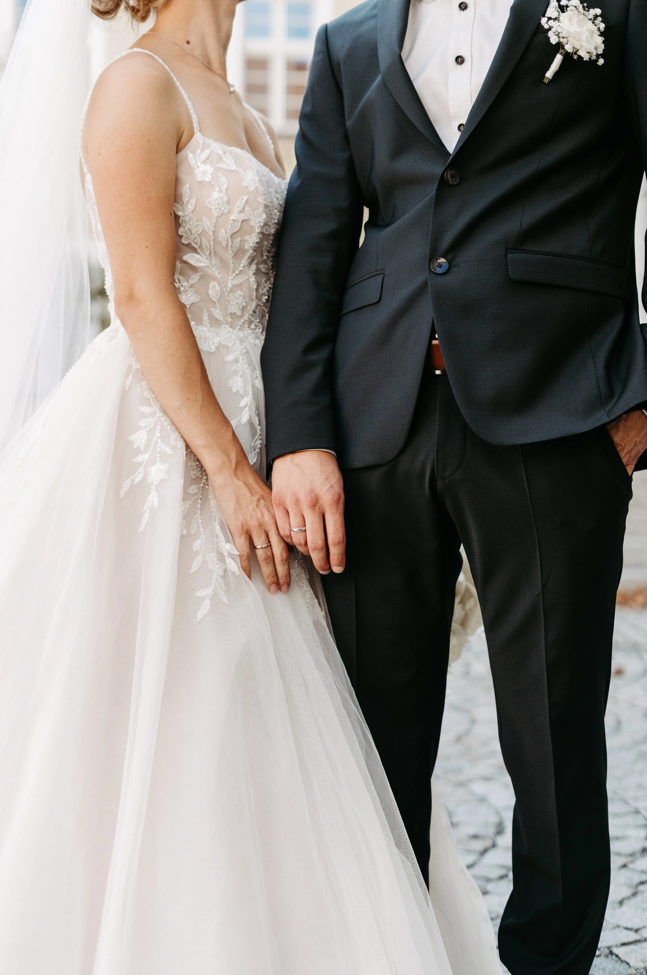 Ein Brautpaar hält Händchen bei einer Hochzeit, die Braut trägt ein weißes, ab Augenschmaus detailliertes Hochzeitskleid, der Bräutigam trägt einen dunklen Anzug.