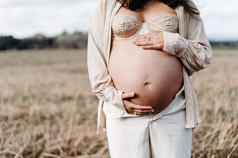 Schwangere Frau steht im Freien, trägt ein offenes Hemd und eine Spitzen-BH, zeigt ihren Babybauch mit einer Hand.