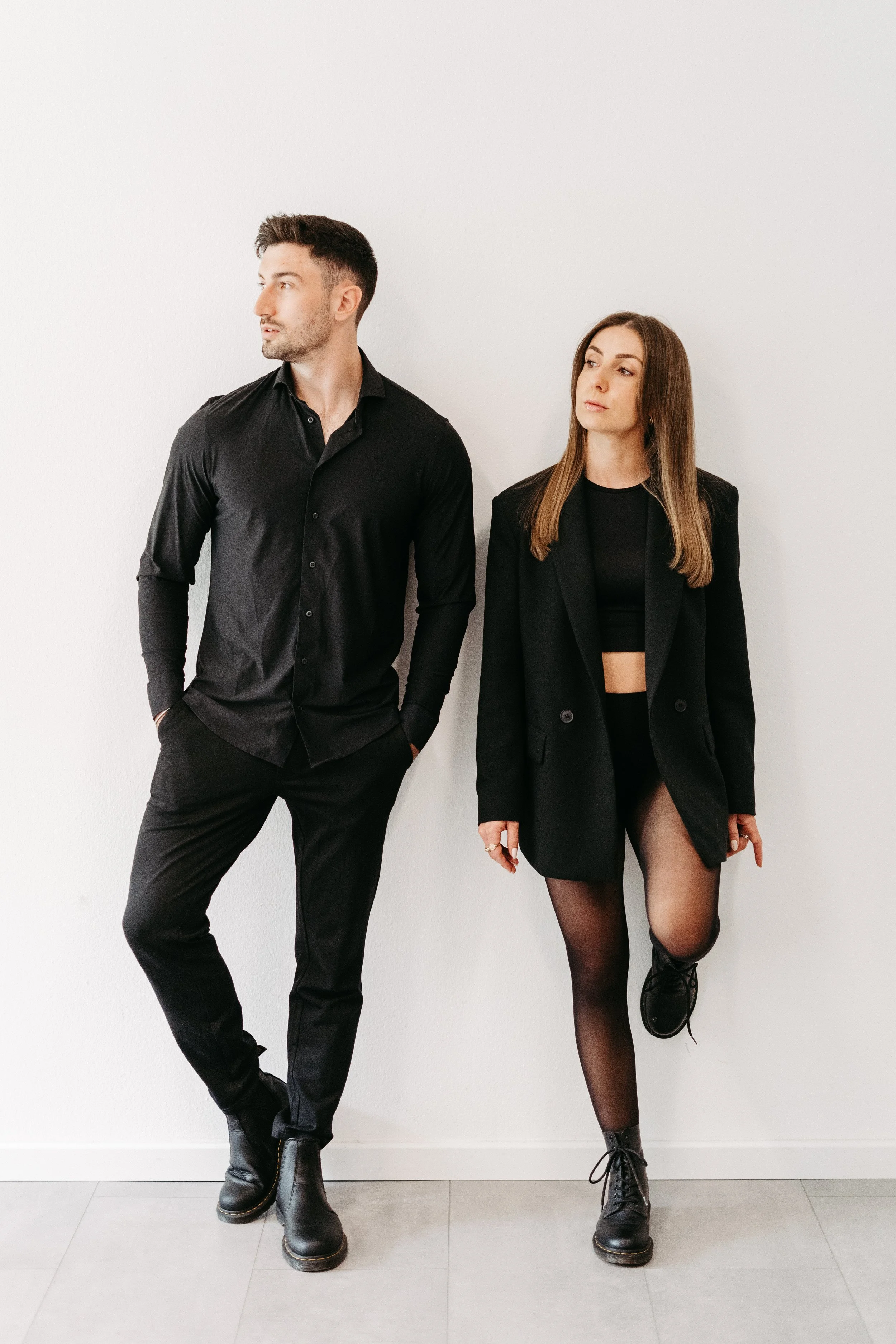 Ein Mann und eine Frau stehen mit schwarzen Kleidern gegen eine weiße Wand, der Mann schaut nach links, die Frau schaut nach vorne