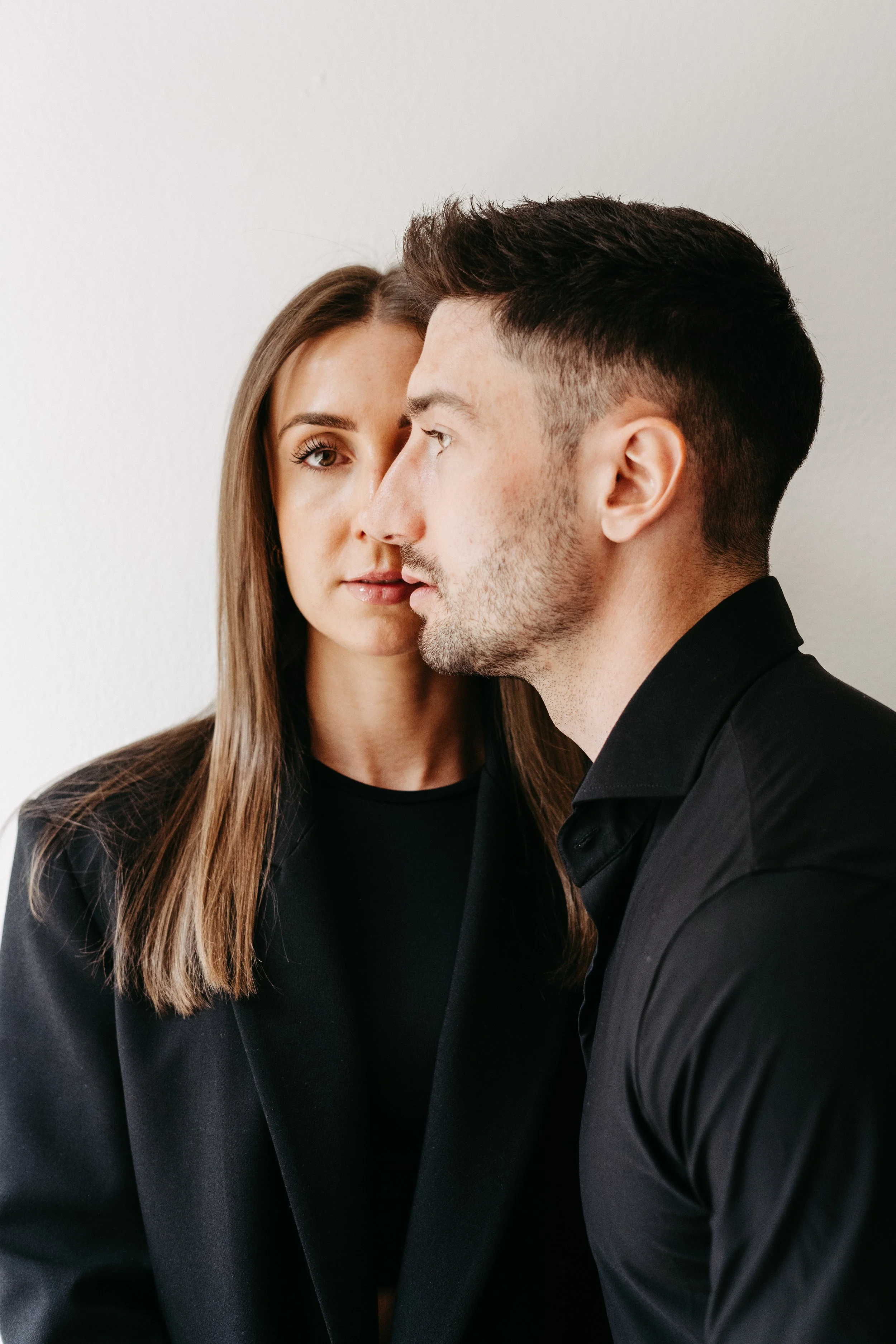 Ein Mann und eine Frau stehen nah beieinander, sehen sich intensiv an und haben schwarze Kleidung an, vor einem weißen Hintergrund.