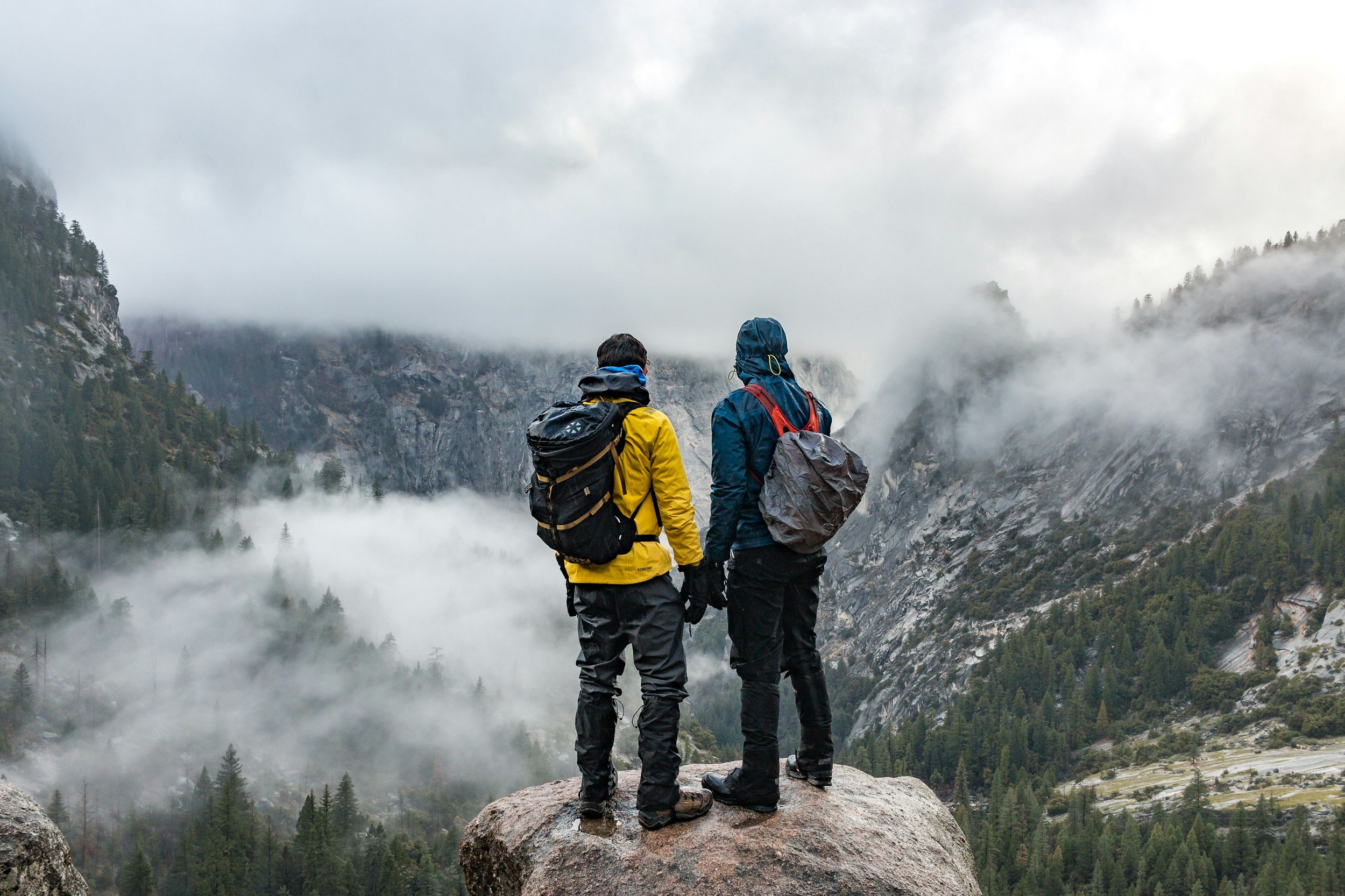 Two hikers standing on a large rock overlooking a foggy mountain landscape with trees, cliffs, and valleys.