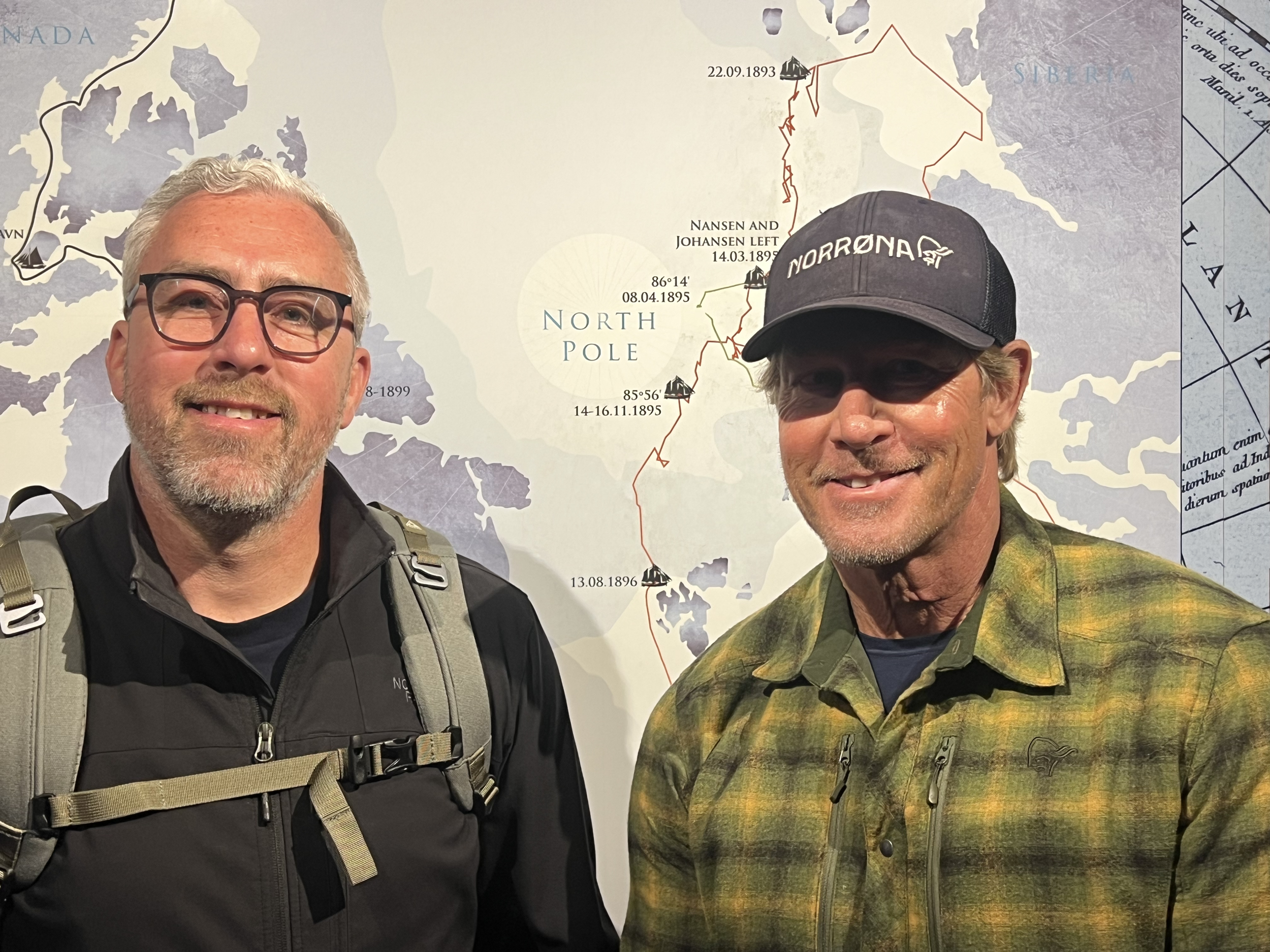 Two men taking a selfie in front of a map of the North Pole, one wearing glasses and a black jacket, the other wearing a cap and a yellow-green jacket.