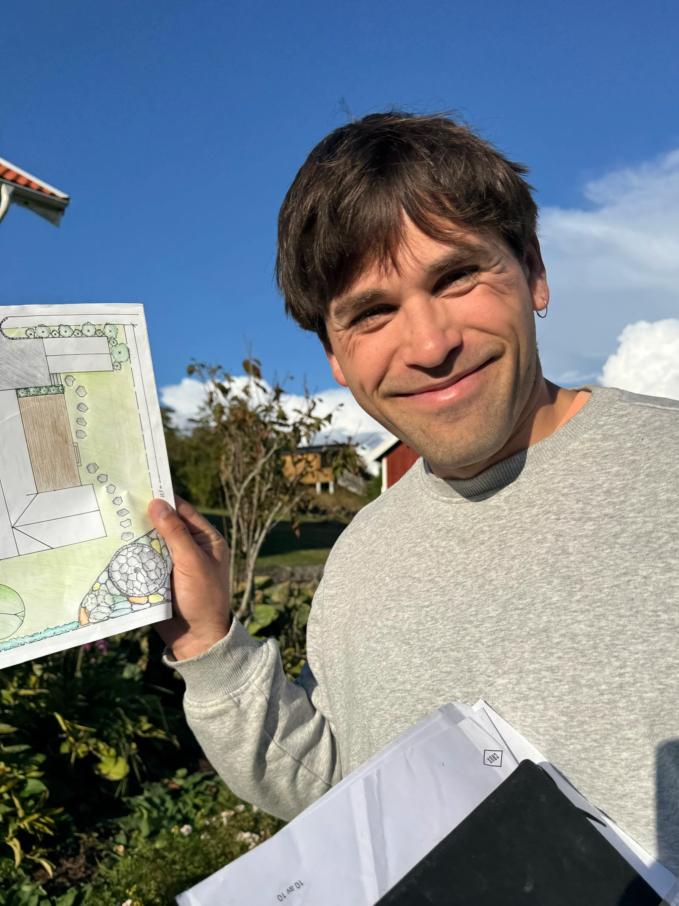 Et smilende menneske holder et arkitekttegning eller plantegning utendørs mot en klar blå himmel.