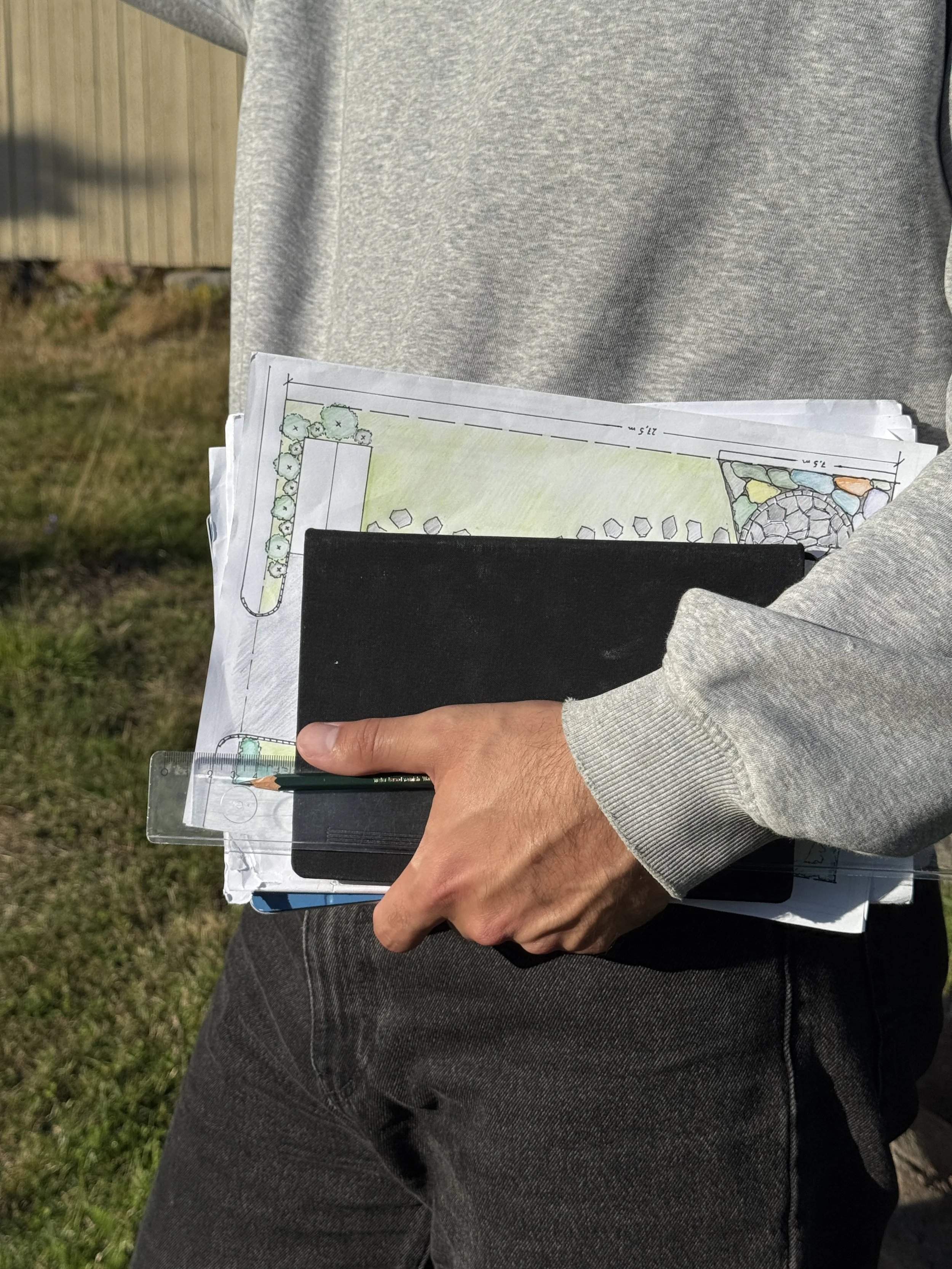 Person holder planer og målebånd foran en bygning, med utendørs bakgrunn.