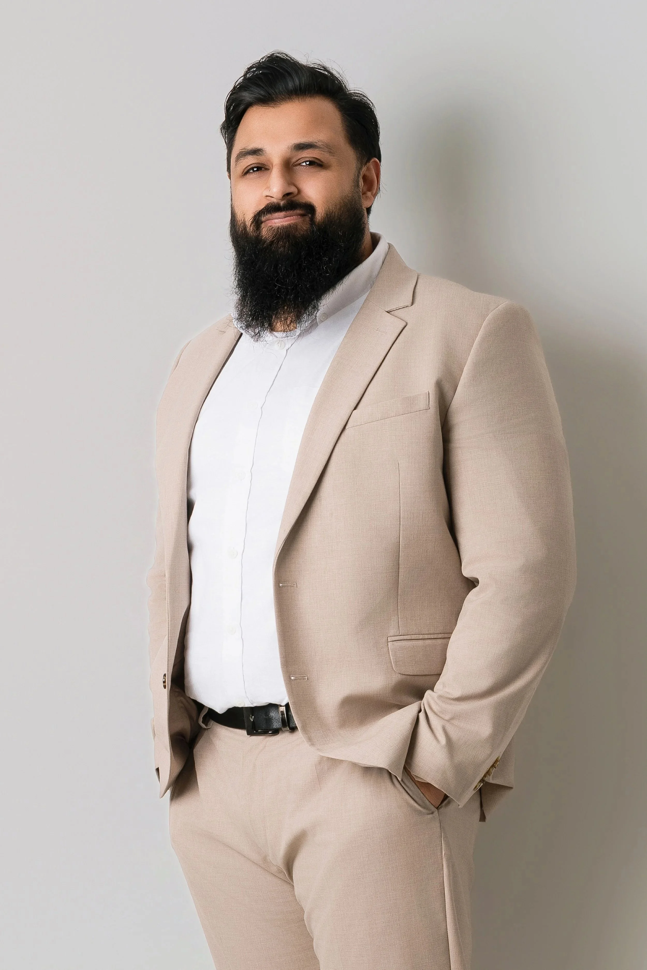 A man with a beard wearing a beige suit and white shirt, standing against a plain light-colored wall.