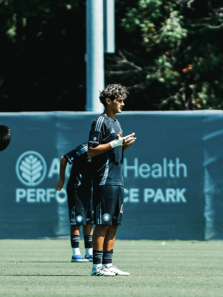 A young male soccer player in black uniform standing on the field during practice or a game, with a teammate behind him, at Art & Health Performance Park.