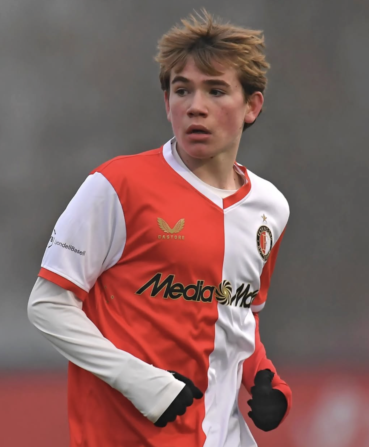 A young male soccer player with tousled light brown hair, wearing a red and white jersey, jogging on a field with a blurred background.