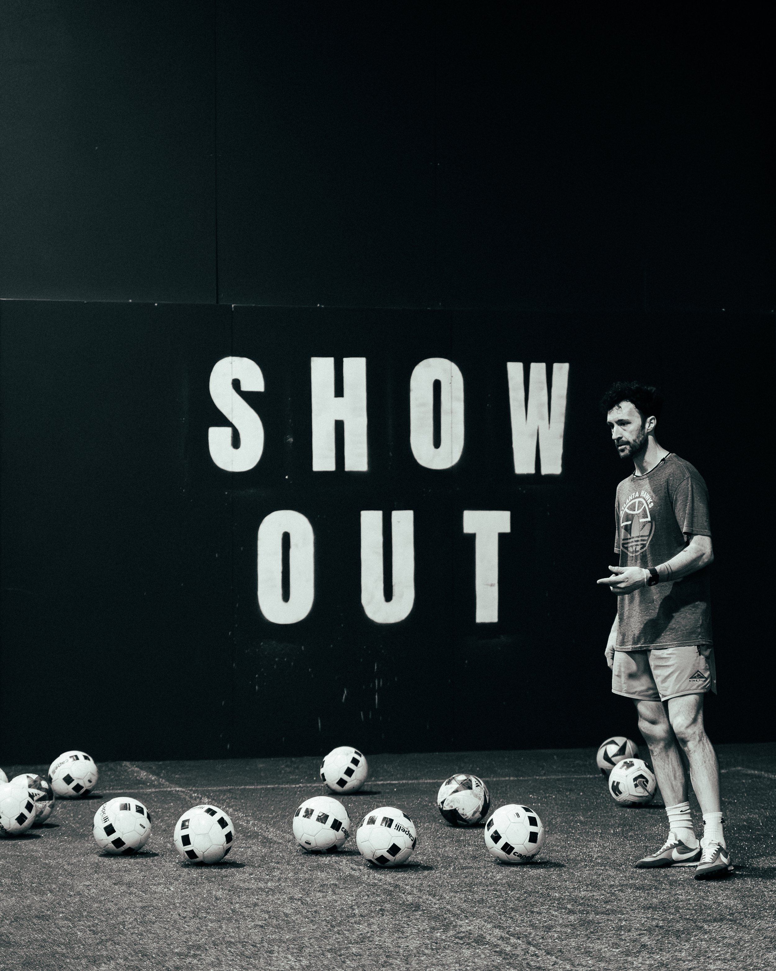 A man standing on an indoor soccer court with multiple soccer balls around him, and a sign in the background that reads 'SHOW OUT'.