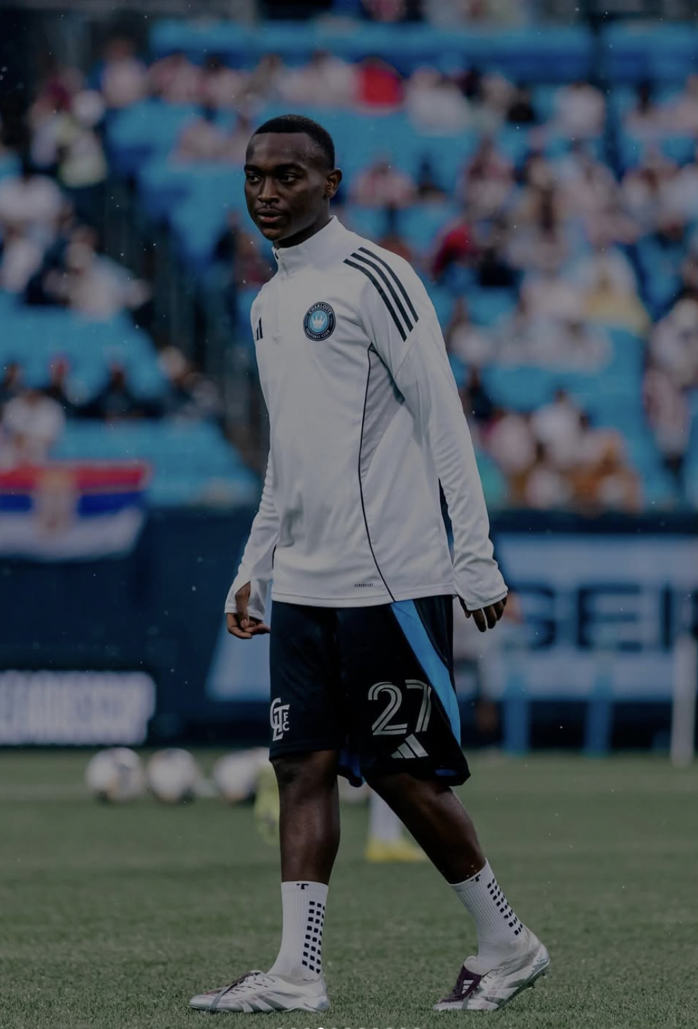 Male soccer player in white and black sportswear standing on a field, with a crowd in the bleachers in the background.