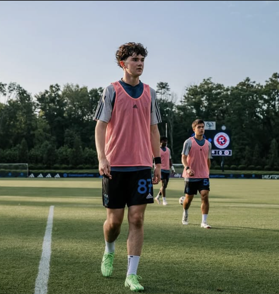 Young male soccer players on a field, wearing gray and pink training bibs, with trees and a scoreboard in the background.