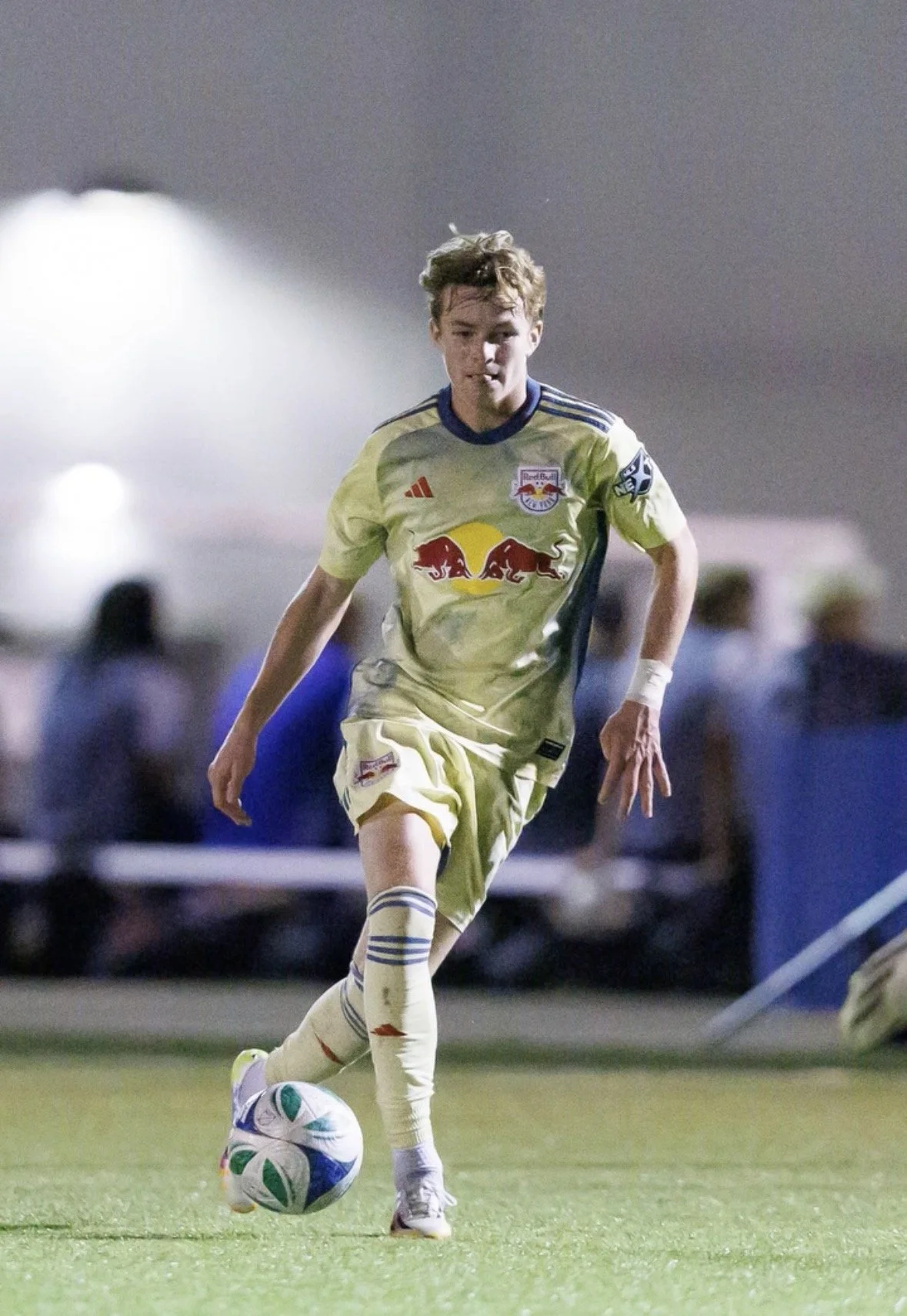 A young male soccer player in a yellow uniform with a Red Bull logo is running on the field with a soccer ball at his feet during a night game, with blurred spectators and lights in the background.