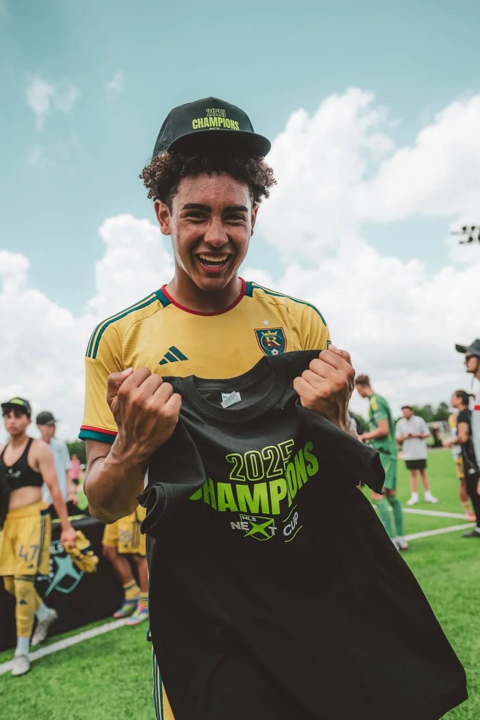 A young man in a yellow soccer jersey holding a black T-shirt that reads '2022 Champions MLS Cup' with a big smile, wearing a black hat that says '2023 Champions,' celebrating on a soccer field.