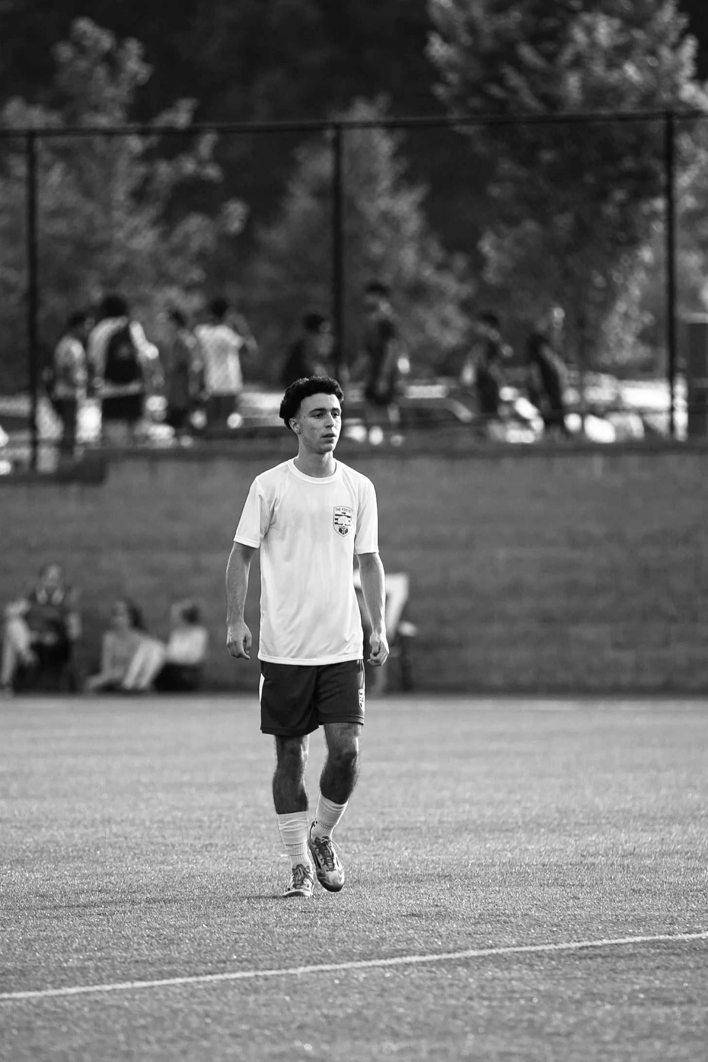 A young man walking on a soccer field during a game, with a fence and people watching in the background, in black and white.
