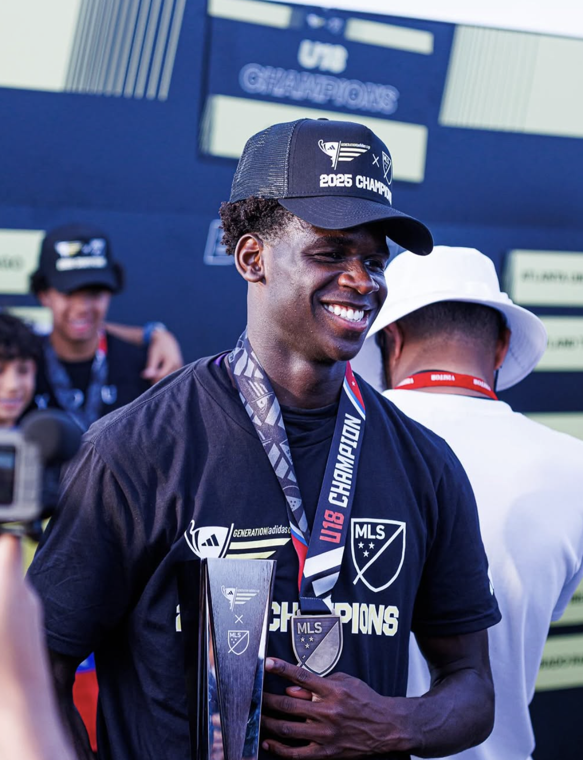 A young man smiling, wearing a black cap and a black t-shirt with 'MLS Champions' and a trophy logo, holding a trophy, with a medal around his neck, celebrating in front of a backdrop with sports branding.