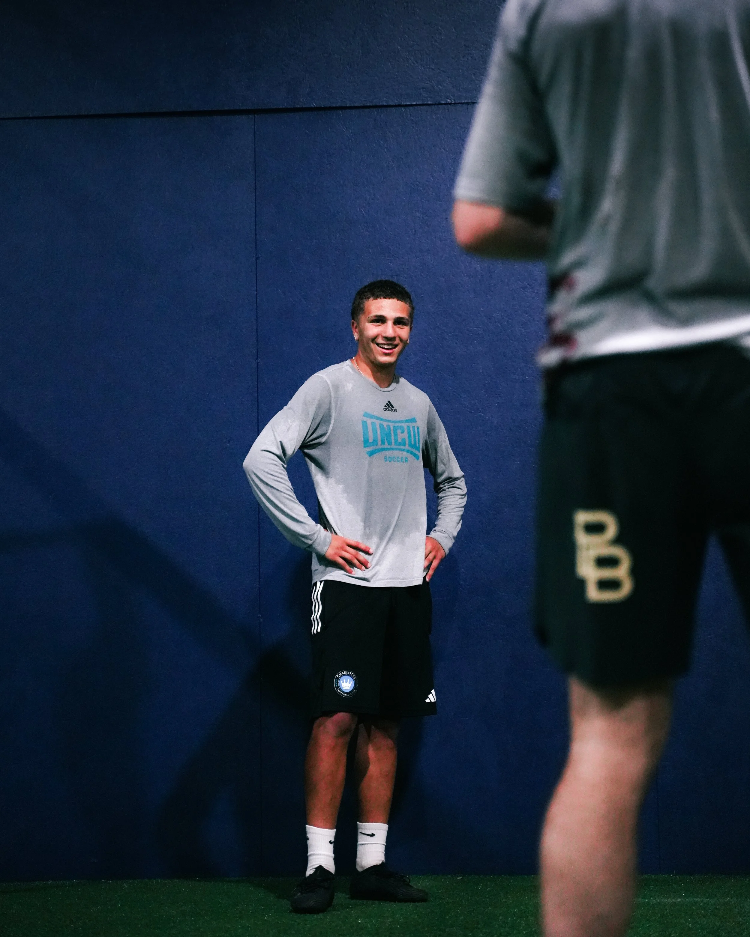 A young man in a gray long sleeve shirt and black shorts standing against a blue wall, smiling and looking at someone in front of him.