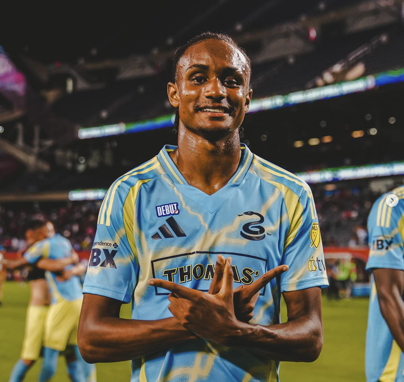 A soccer player in a blue and yellow jersey standing on a field, making a gesture with his hands, with other players in similar uniforms in the background.
